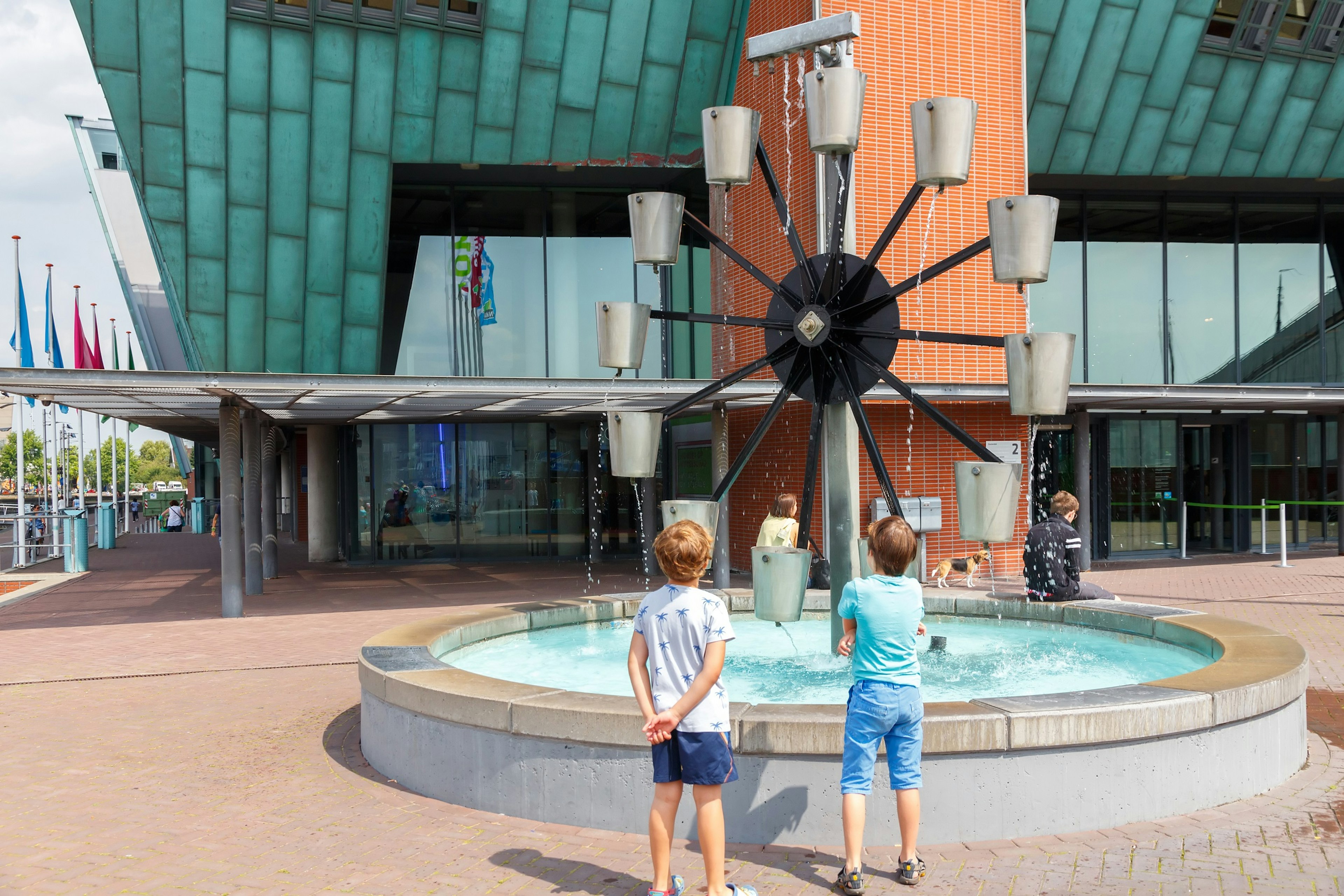 Two children stand watching a water wheel where cups of water are lifted as the wheel rotates