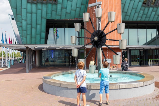 Two children stand watching a water wheel where cups of water are lifted as the wheel rotates