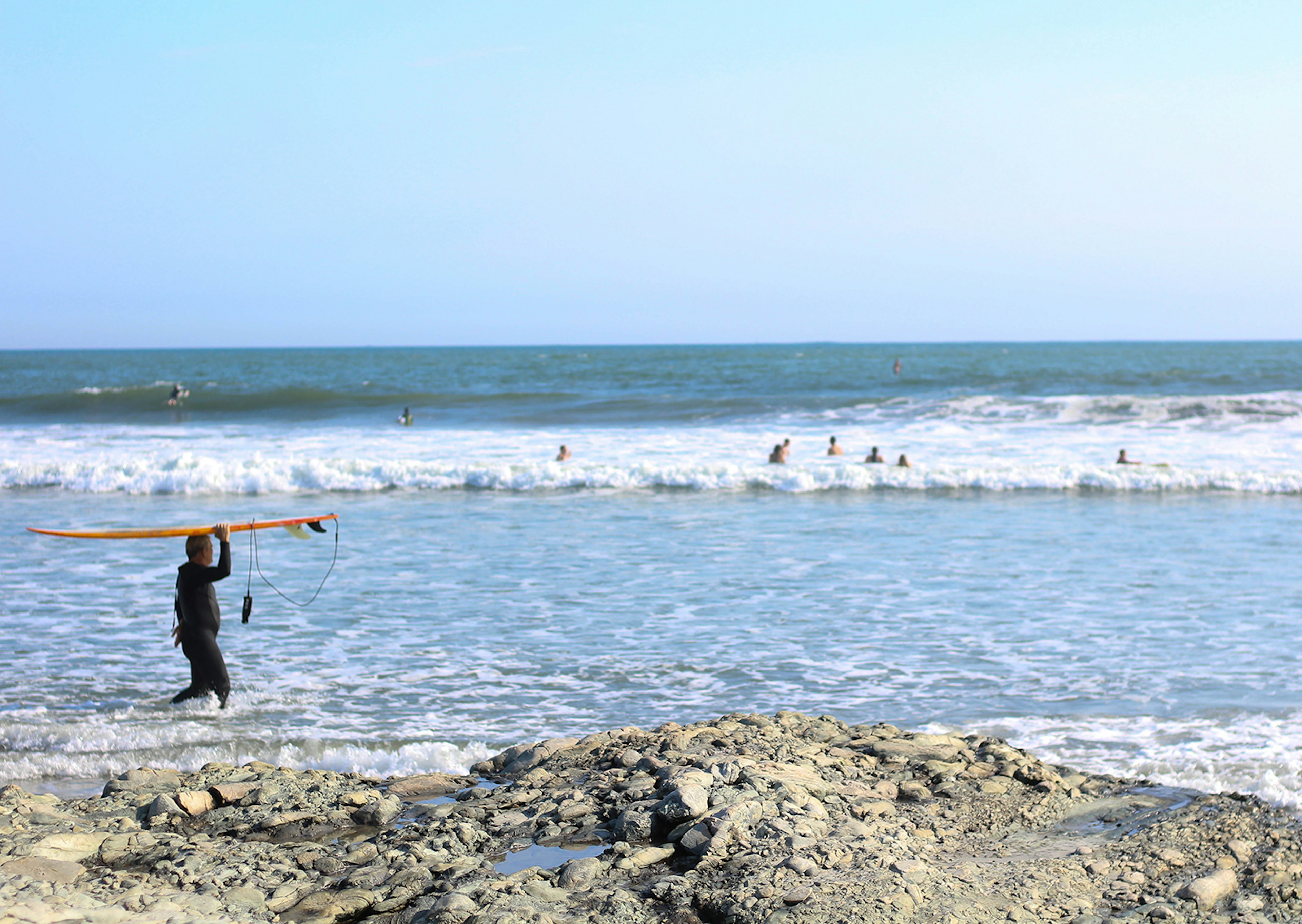 Long shot of a lone surfer carrying a board above their head on a rocky Rhode Island beach, with waves in the background © Anna Saxon / Lonely Planet