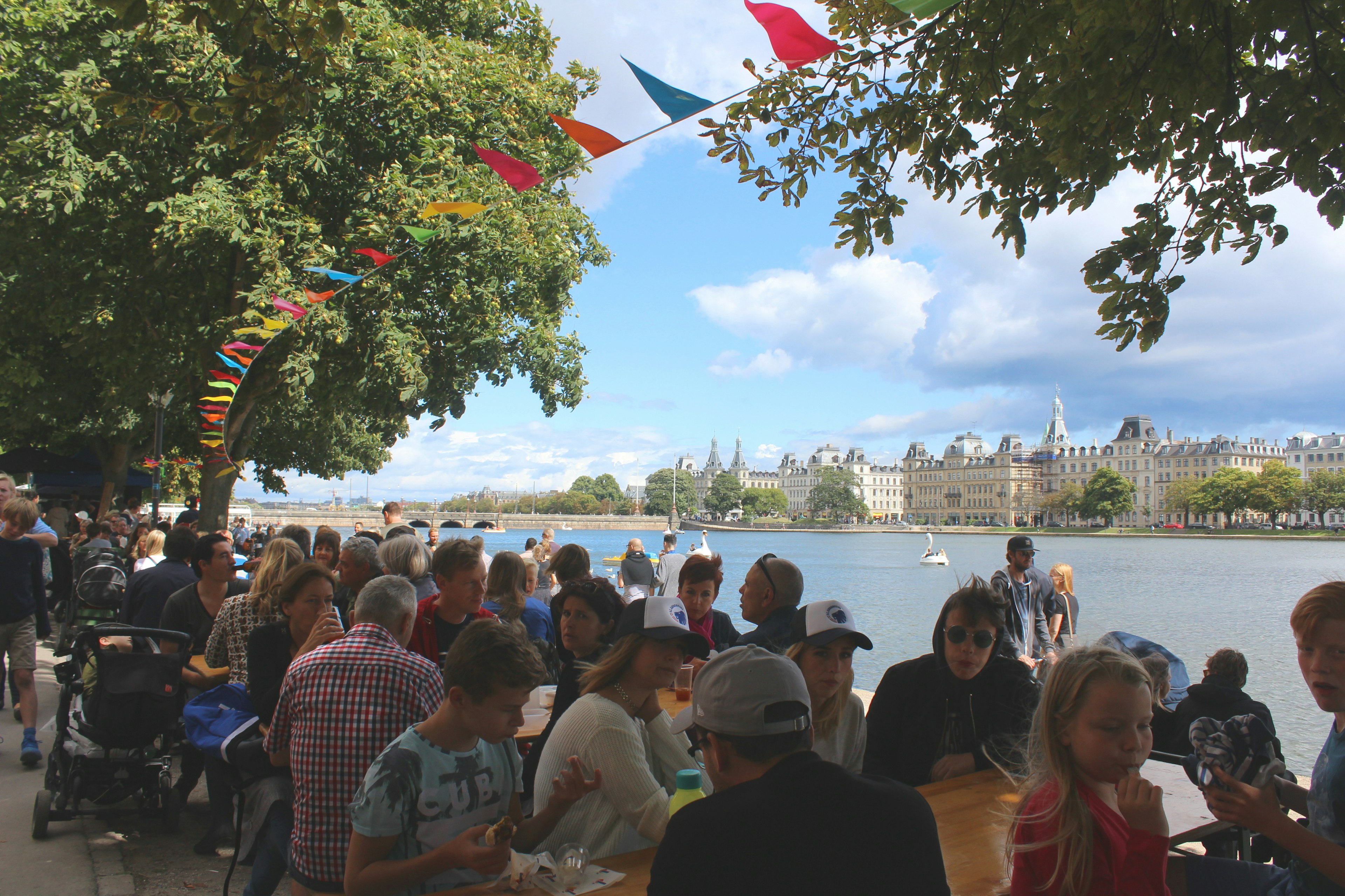 People sit at picnic tables along the banks of the Copenhagen lakes in ø with colourful bunting and leafy trees overhead. There are pedalos and a white-stone palace in the background.