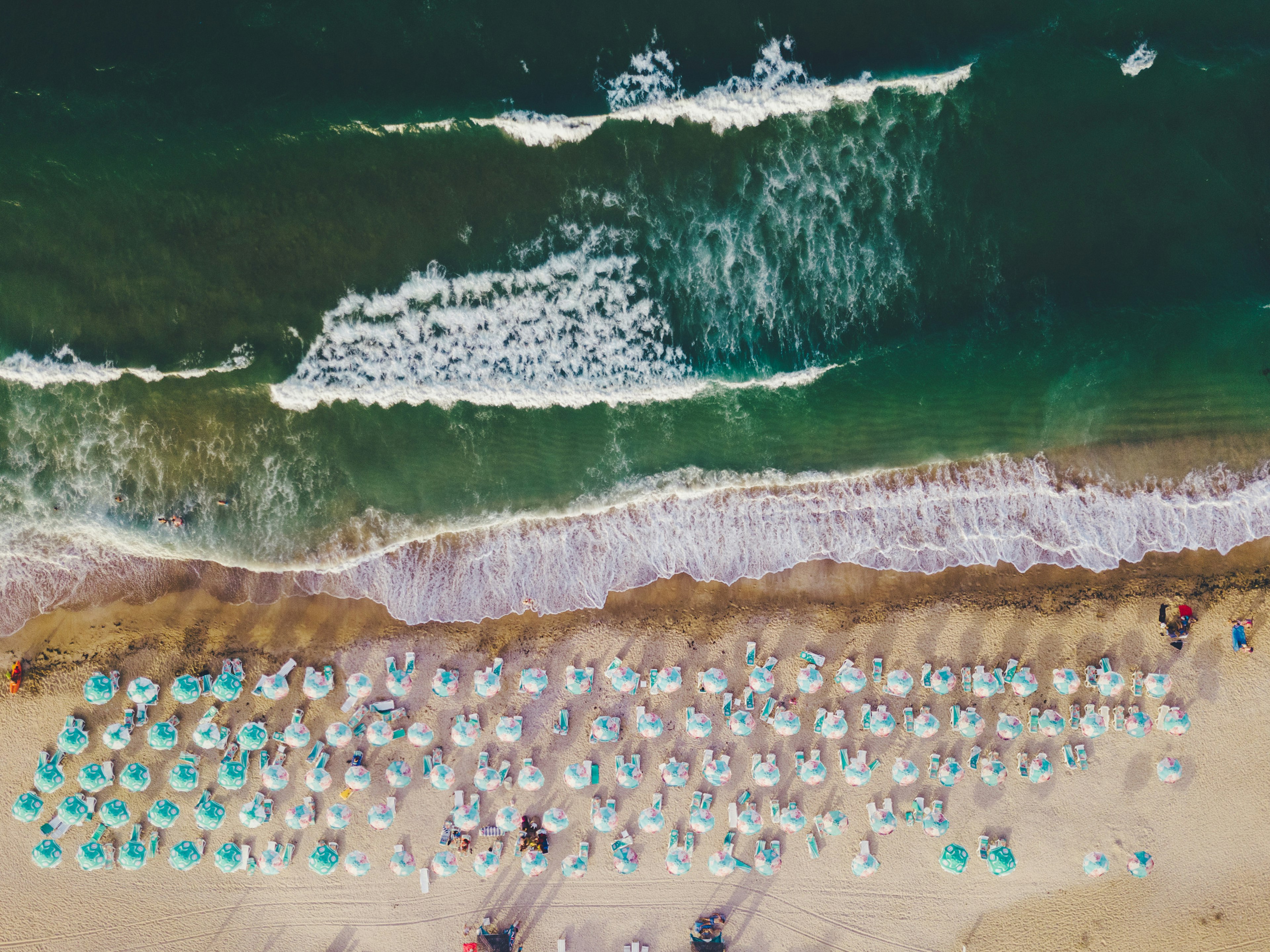 An aerial view shows bright umbrellas on the beach and a deep ocean in front.