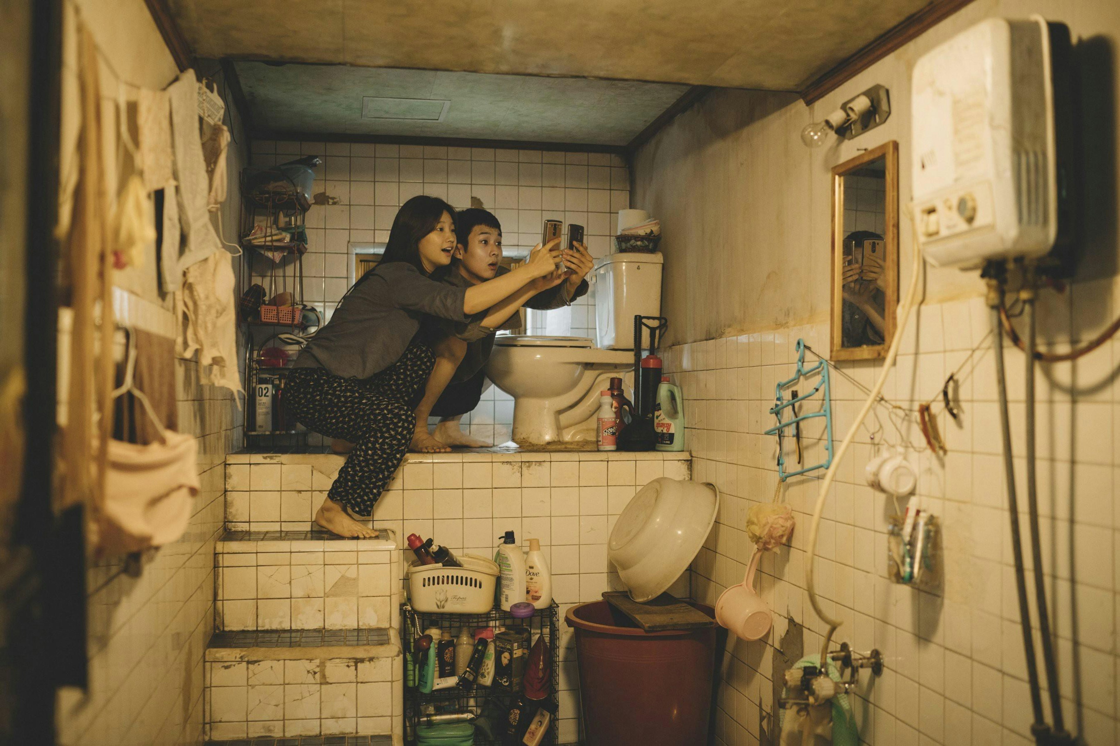 Actors Park So-dam and Choi Woo-sik in a still from 'Parasite': they are taking selfies, perched on steps beside a toilet in a dishevelled, tiled bathroom.