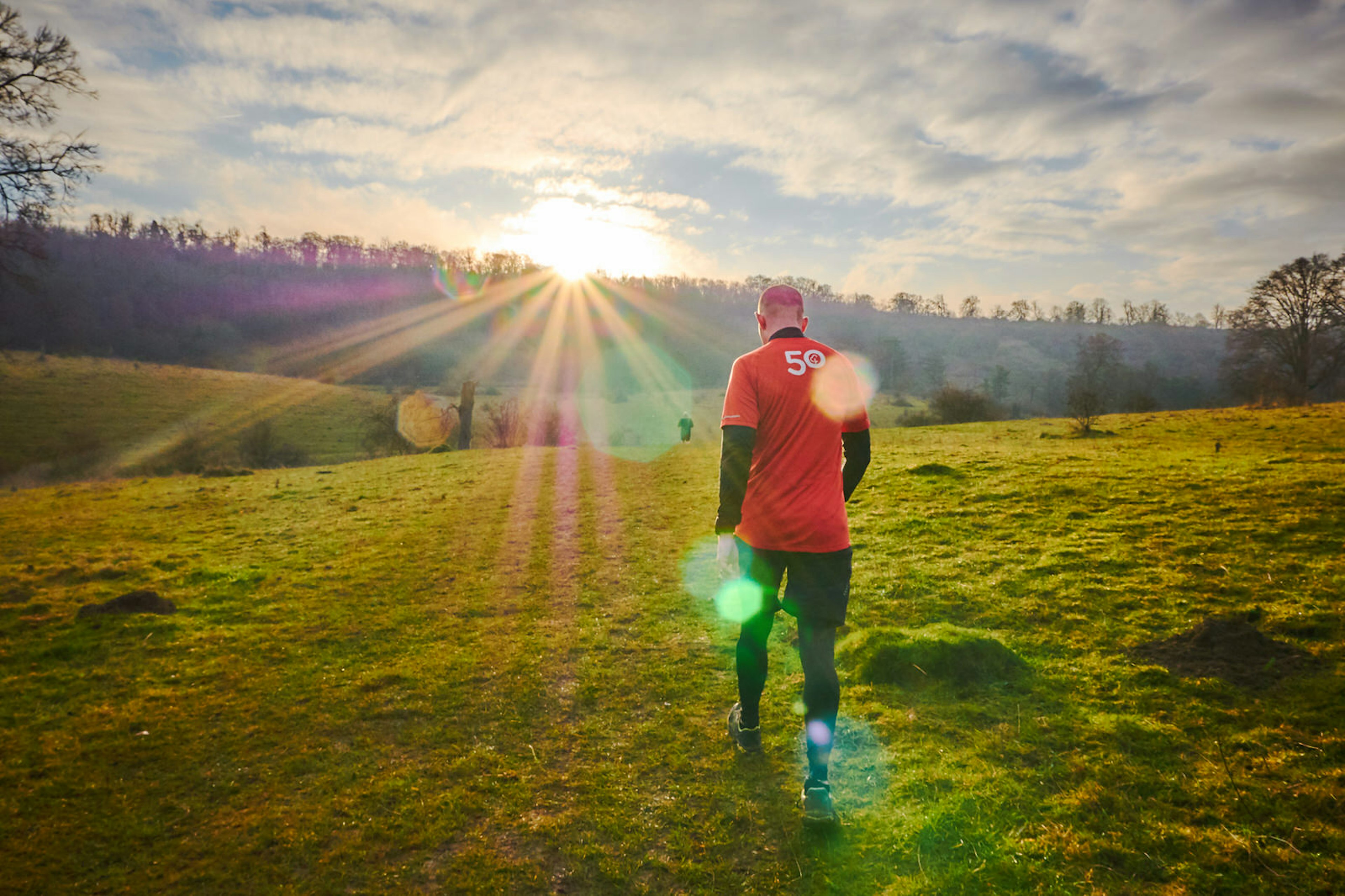 A man walks across the top of a grassy hill as the sun rises above trees on the horizon, causing rays and stars of light to fill the picture