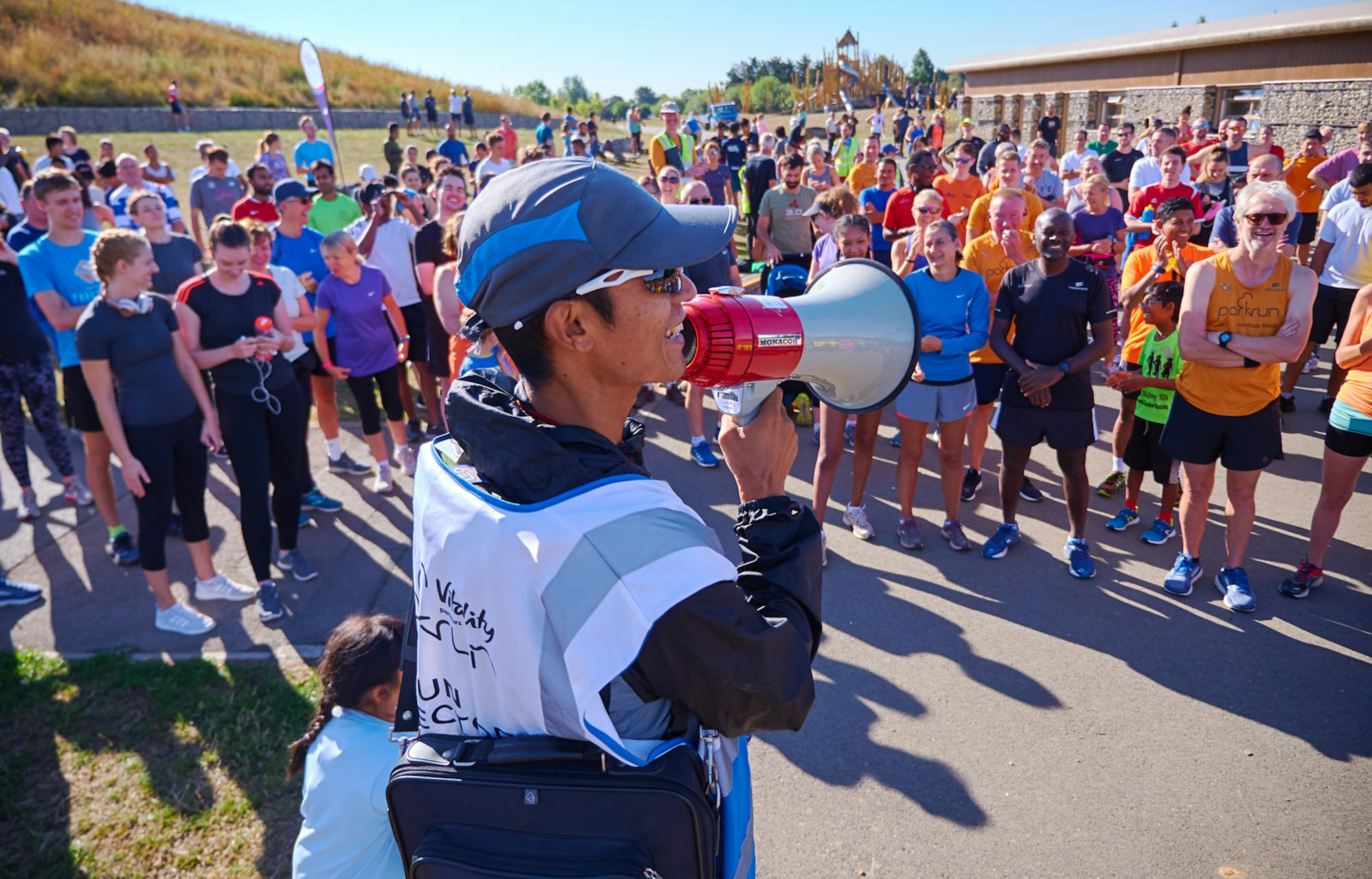 A man in blue baseball cap and sunglasses speaks into a megaphone to a crowd of runners in colourful kit