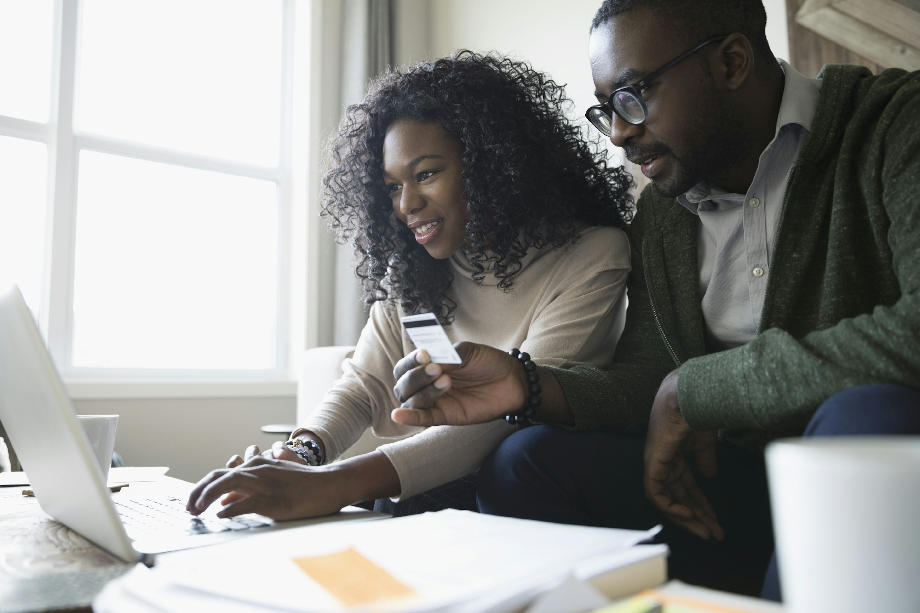 A couple sits beside each other on the couch. The woman is using the computer and the man is holding a credit card.