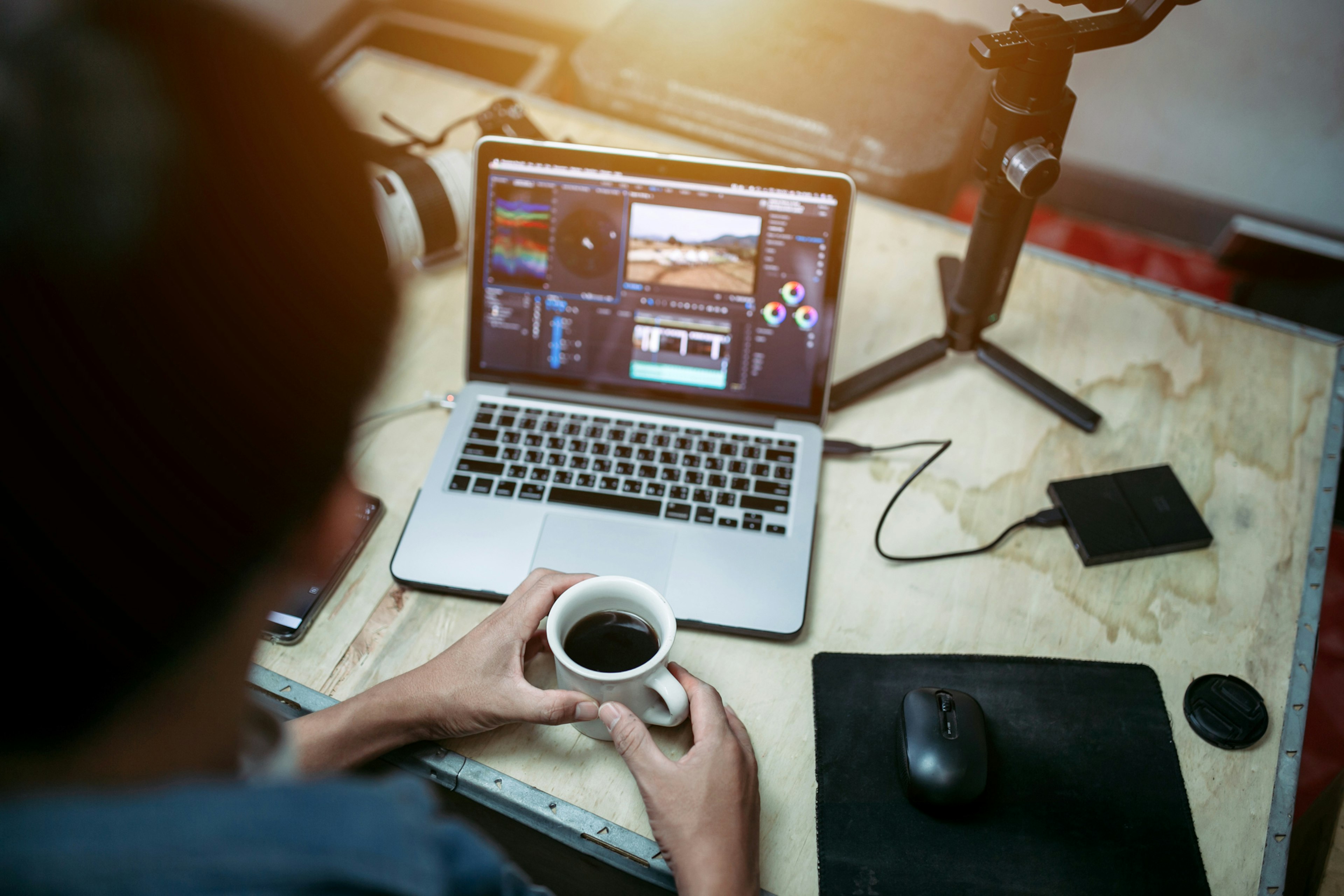 A person with dark hair in a dark blue denim shirt sits in front of a laptop with photo editing software on the screen, an external hard drive, and other photo backup equipment