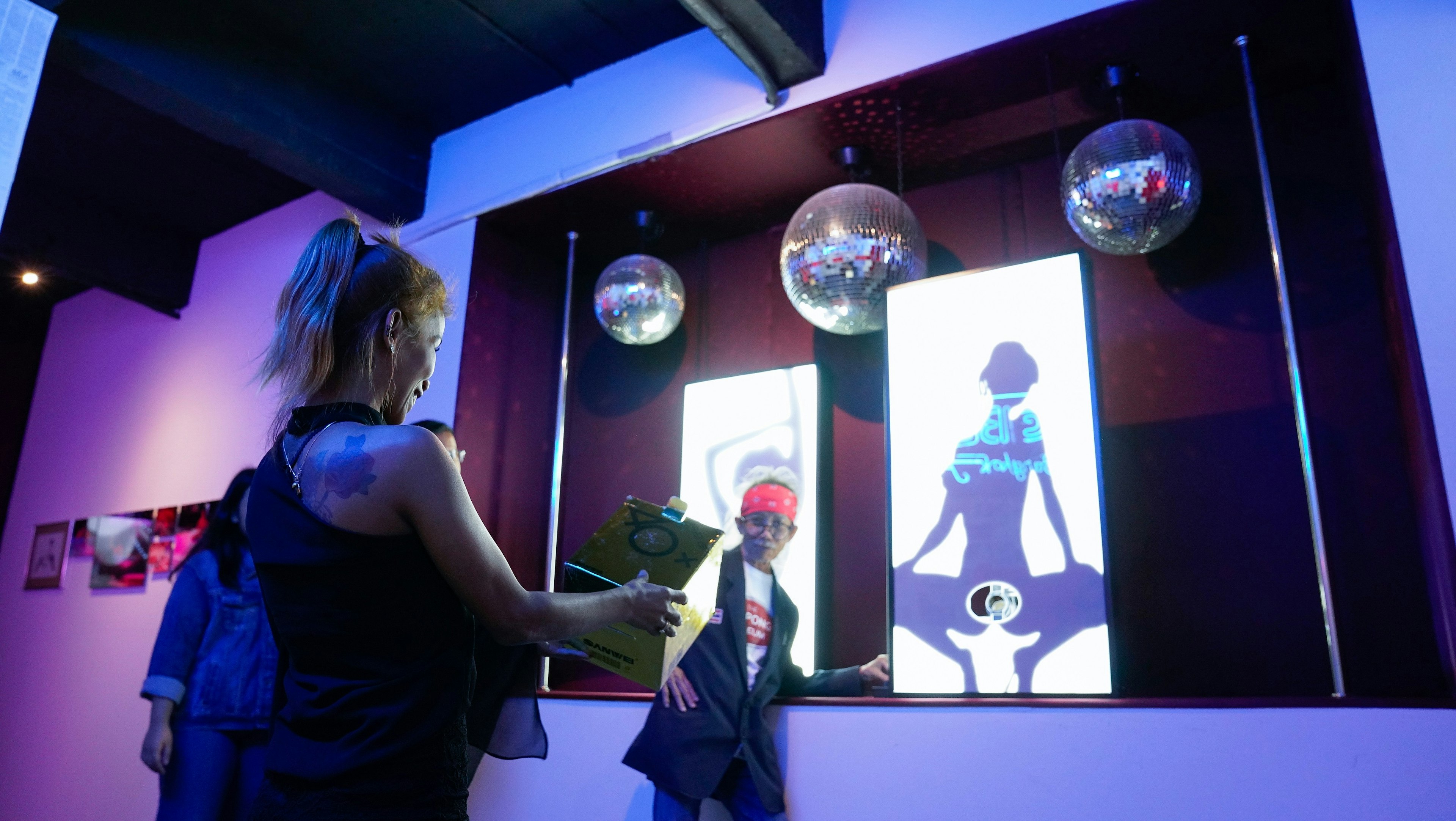 A woman stands with a box waiting to catch a ping pong ball that will be shot from a hole, situated between the legs of a silhouette of a lady.