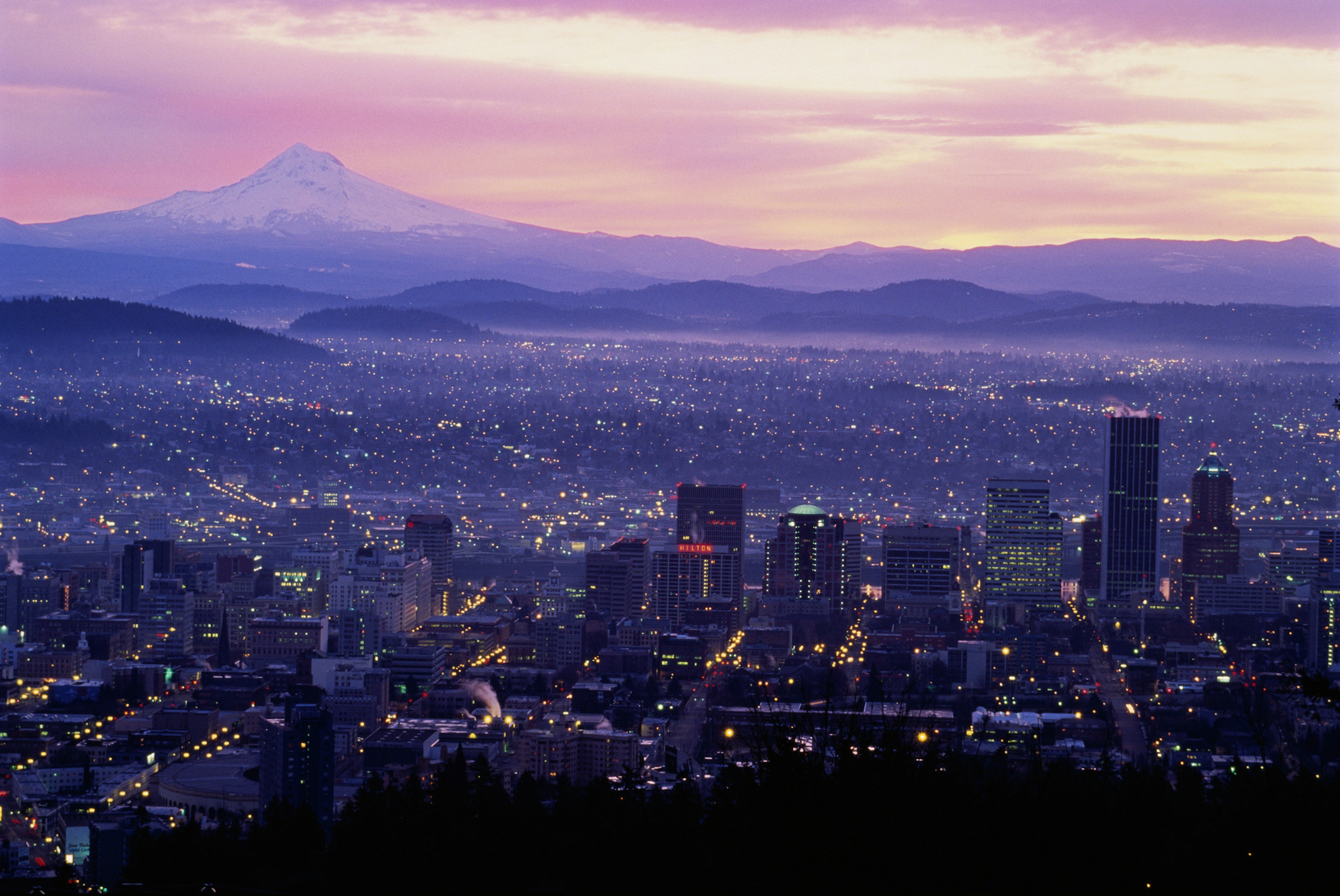 The sun rises over Portland, Oregon, washing the city's downtown skyscrapers and suburban sprawl in lavender and pink light as Mt. Hood rises over the landscape in the top left of the frame