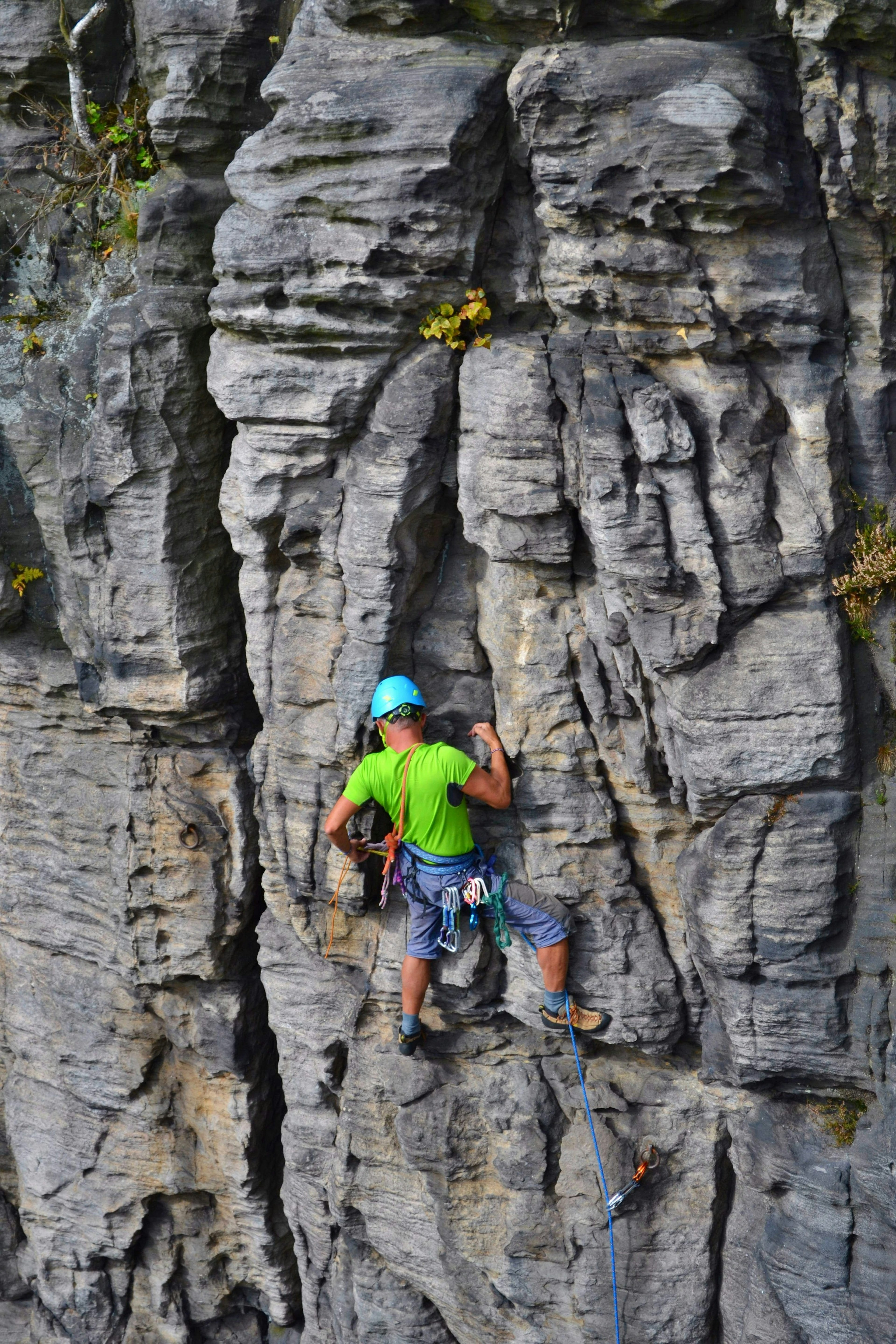 A climber in a bright green t-shirt and sky-blue helmet clings to a rock face that is full of cracks and crevices.