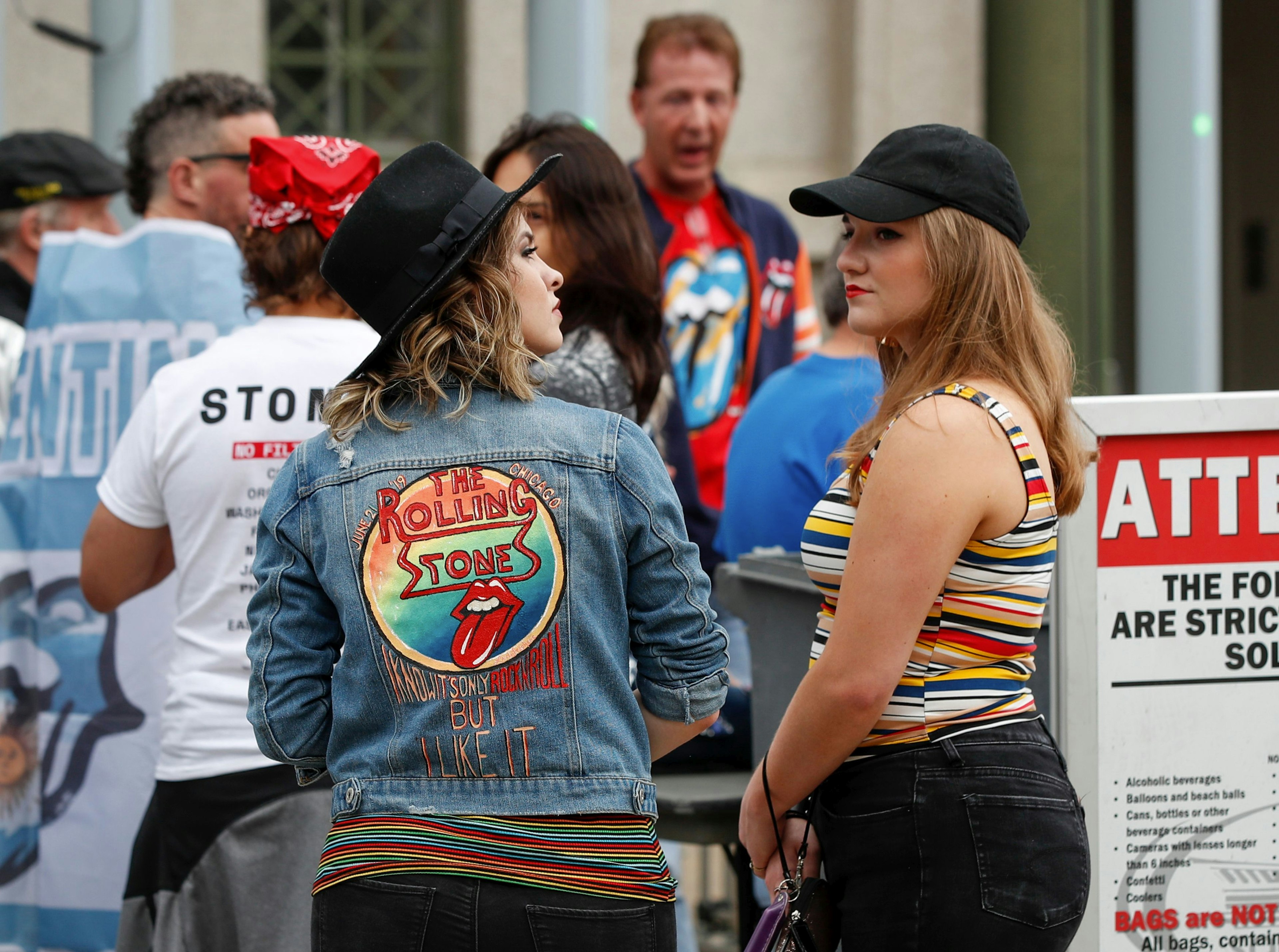 A pair of women in black jeans and black hats stand outside Soldier Field in Chicago ahead of a Rolling Stones concert. One has her back to the camera and is wearing a denim jacket embroidered with the Rolling Stones tongue logo and the lyric