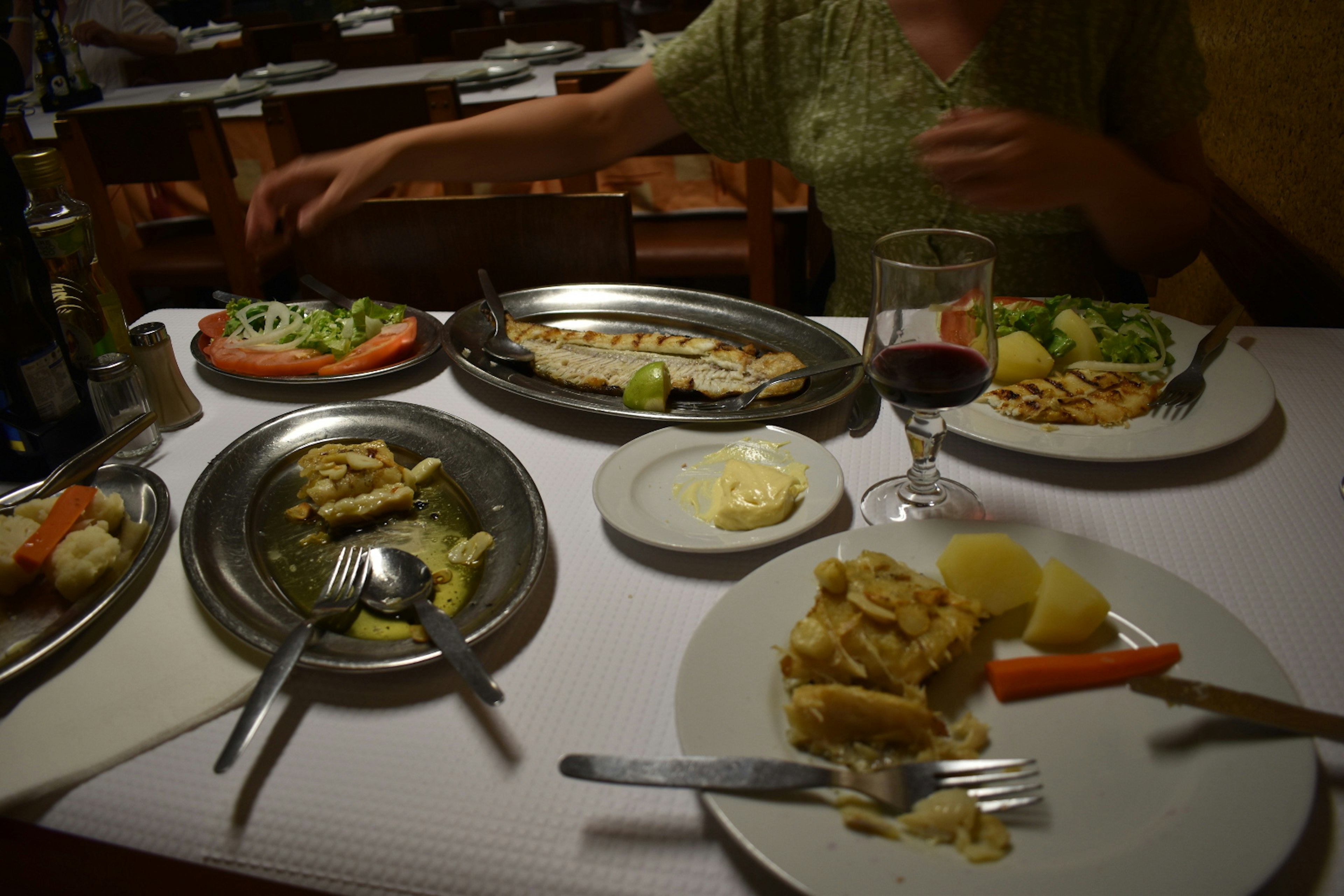 Half-eaten dishes of fish lie around a table with glasses of red wine.