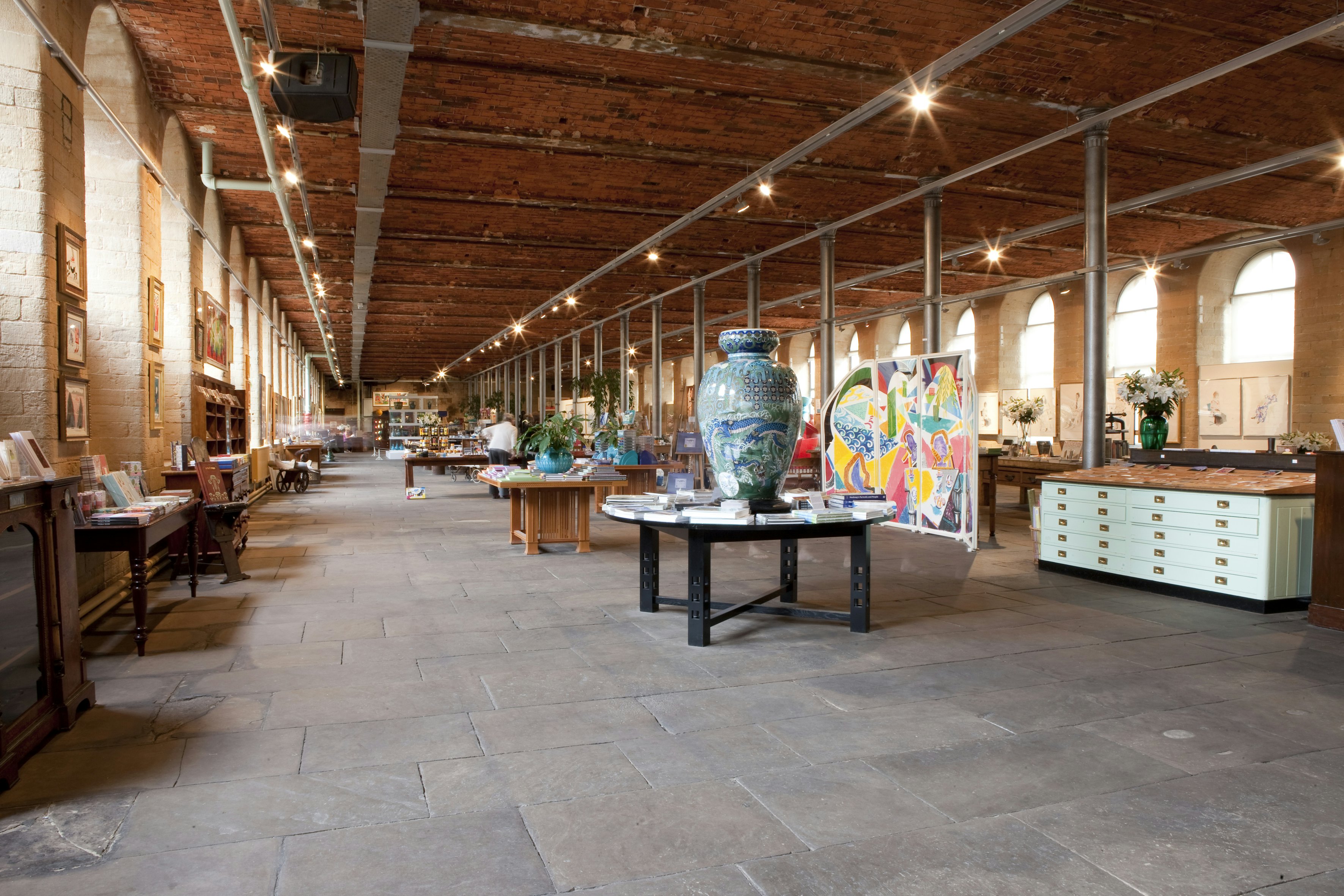 The minimalist interior of a vast, long room at Salts Mill in Saltire; the room has floor-to-ceiling arched windows running each side and is filled with tables displaying artworks, books and other items.