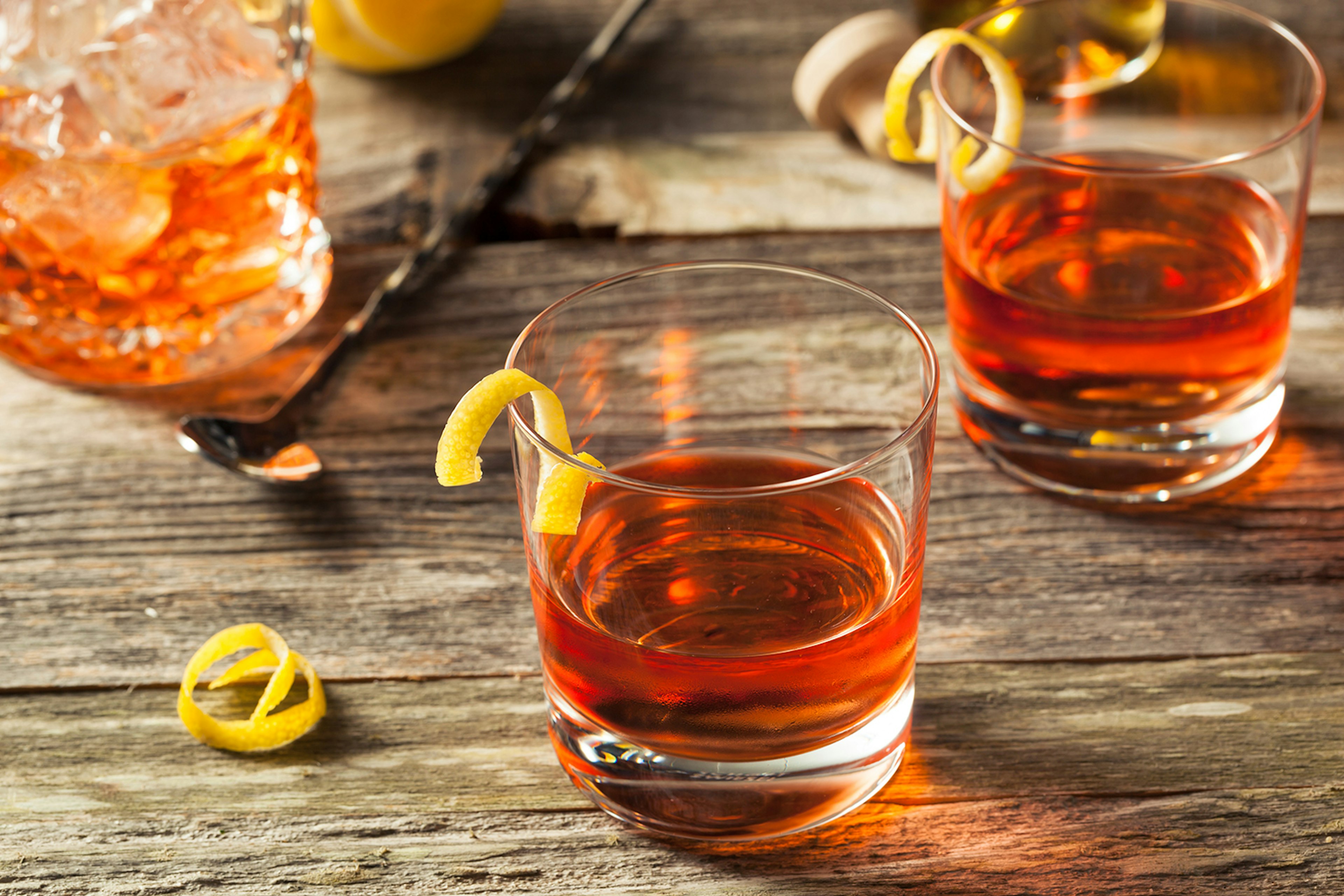 side shot of two rocks glasses with lemon peel garnishes on a wooden table – each glass contains a Sazerac cocktail