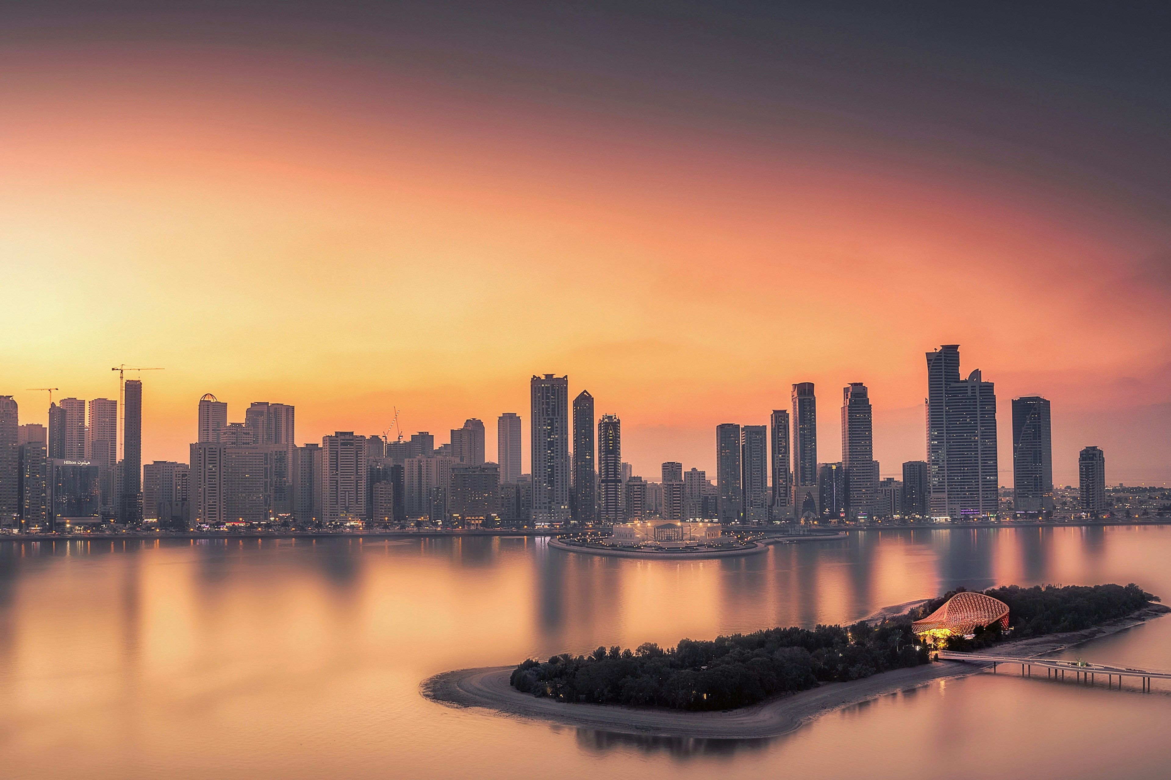 The skyline of Sharjah, comprising numerous skyscrapers, juts up into a bright orange sky at sunset; in the foreground is the forest-covered island of Al Noor, which has some dazzling architecture at its centre.