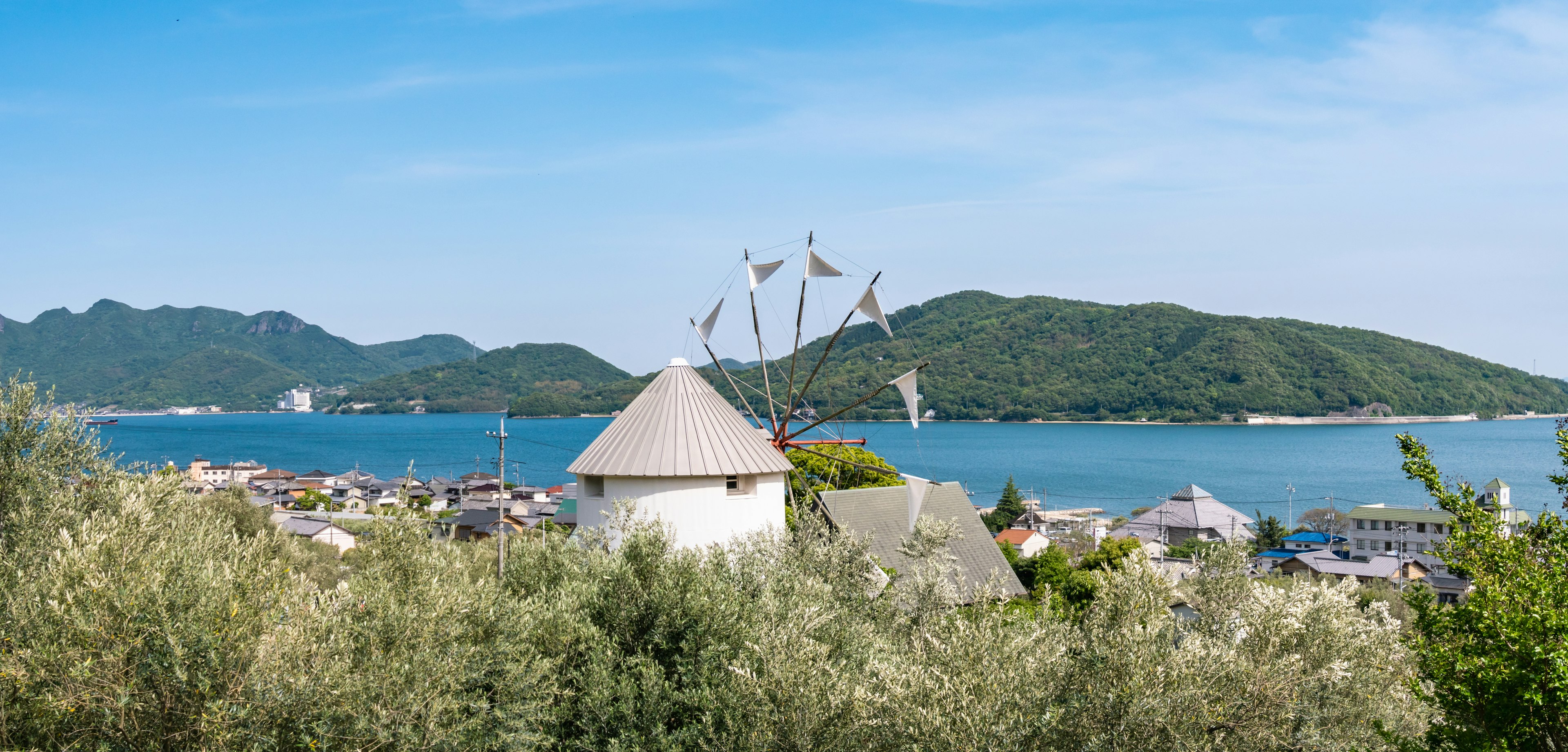 A white windmill with flag-like silver blades and a round silver conical roof overlooks the ocean and green forested islands across the bay. In the foreground are the olive groves of Shdoshima in Kagawa Prefecture, the first place to cultivate olives in Japan