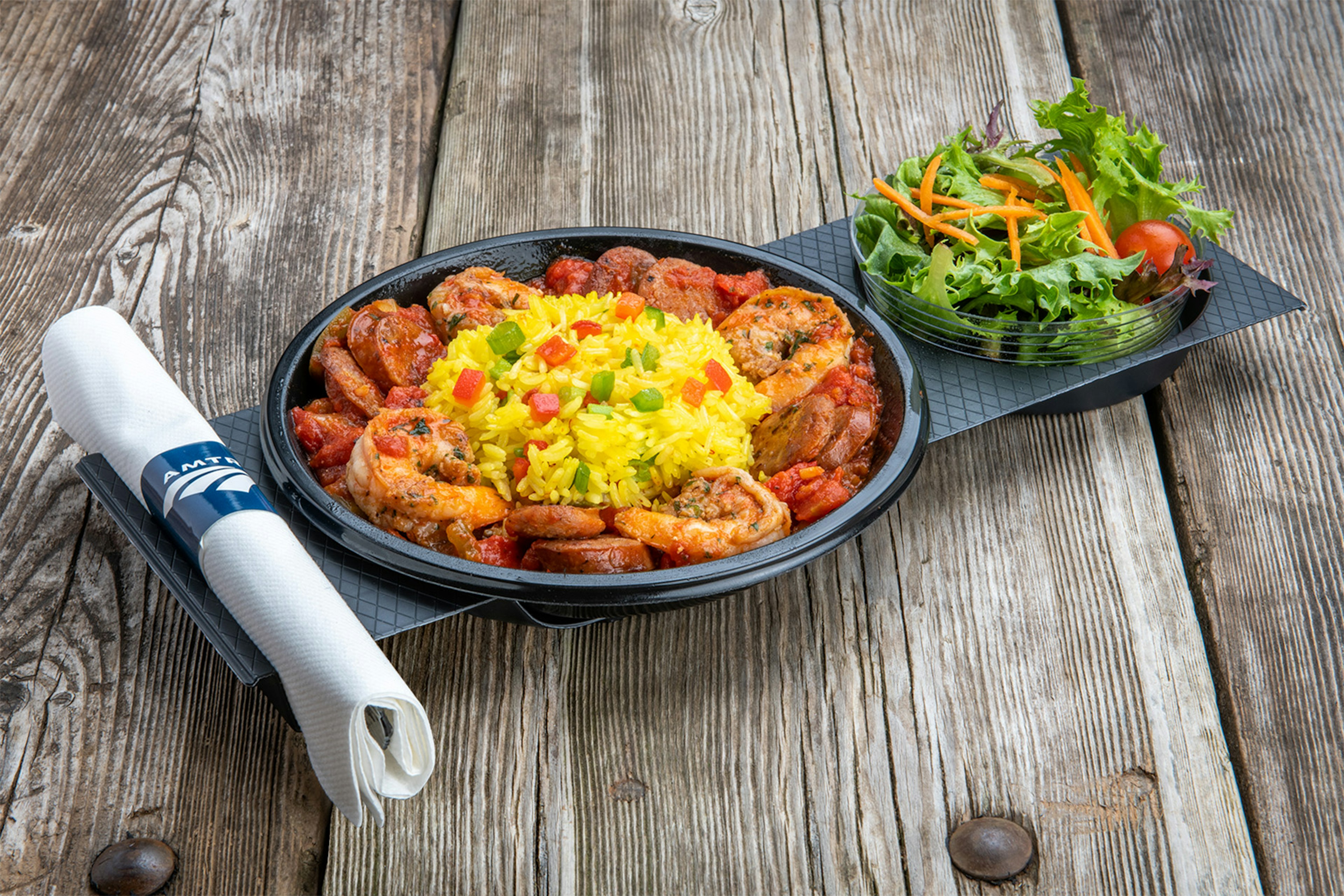 A product shot of the new pre-prepared meals that will be available on certain Amtrak routes. It's a boal of shrimp in a red tomato sauce with a round portion of yellow rice studded with green adn red bell peppers, in a black plastic bowl on a black plastic tray. A small plastic cup of a fresh garden salad topped with shredded carrots and a round cherry tomato is on the side. A paper napkin with silverware inside is wrapped with a navy paper band with the Amtrak logo. The meal sits on rough-hewn wood.