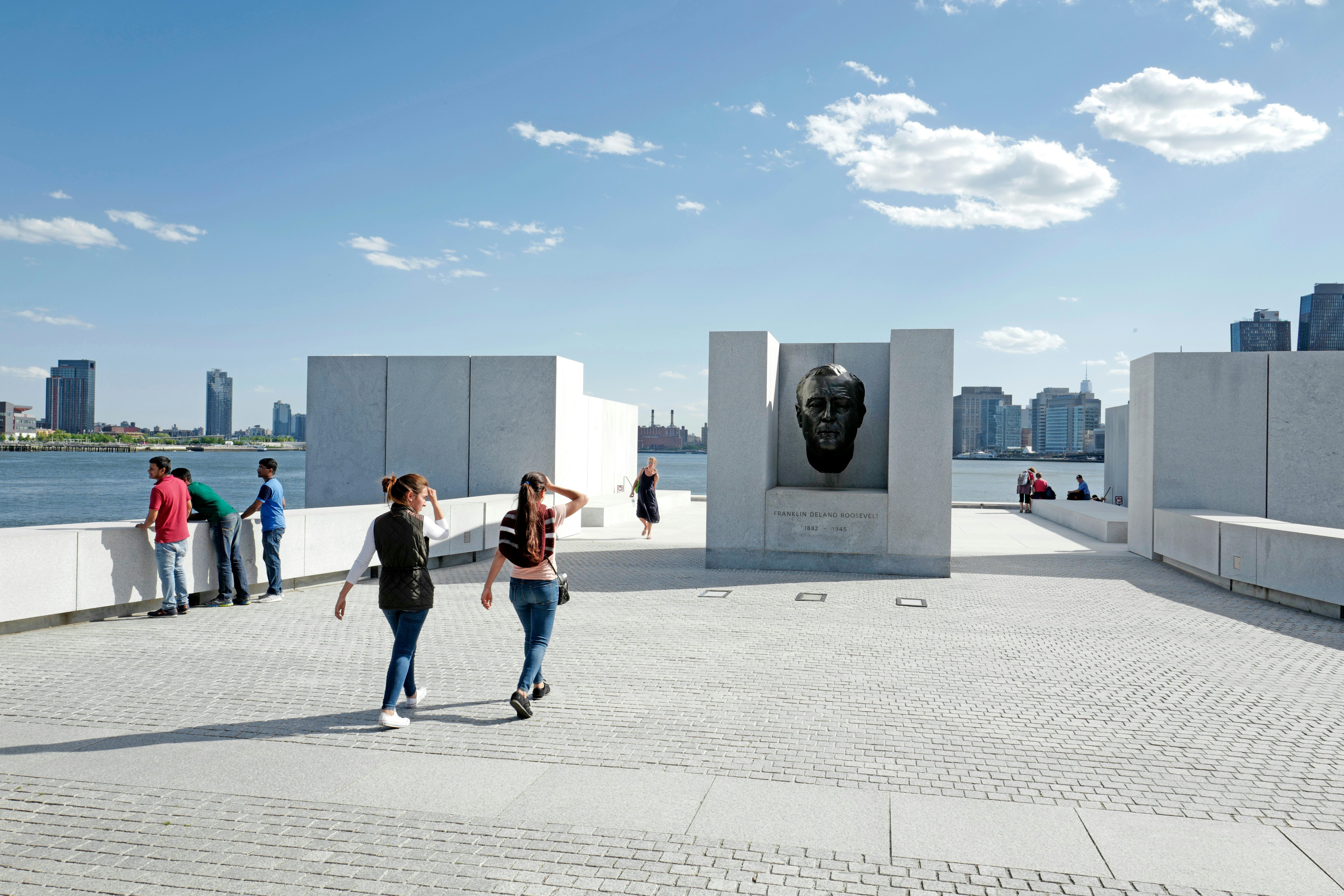 People walk on an open plaza of white bricks toward a dark bust in a white stone block; a river is in the background.