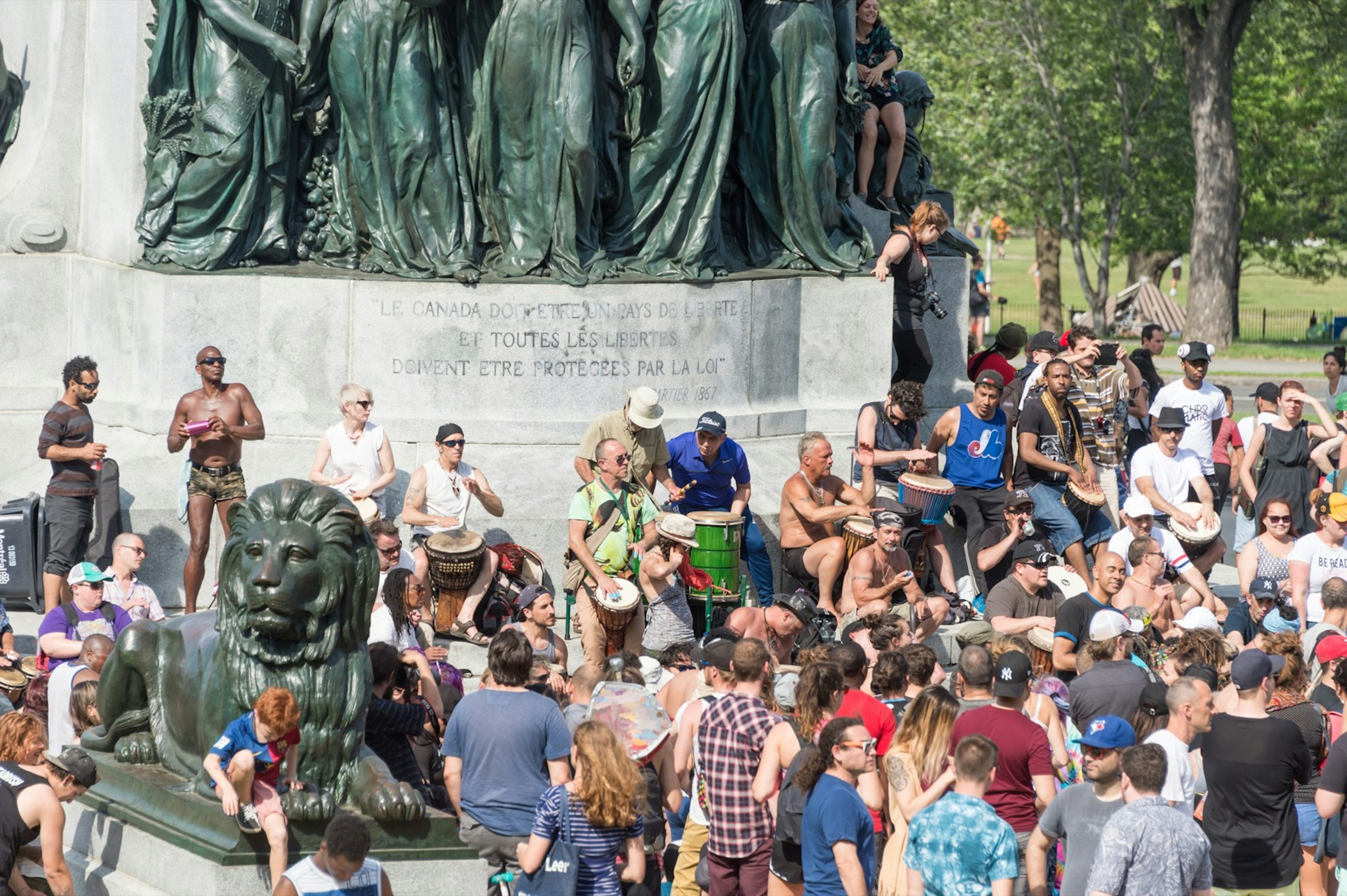 A large group of people play tam-tams and other drums as other people dance and revel in the nice weather around a statue