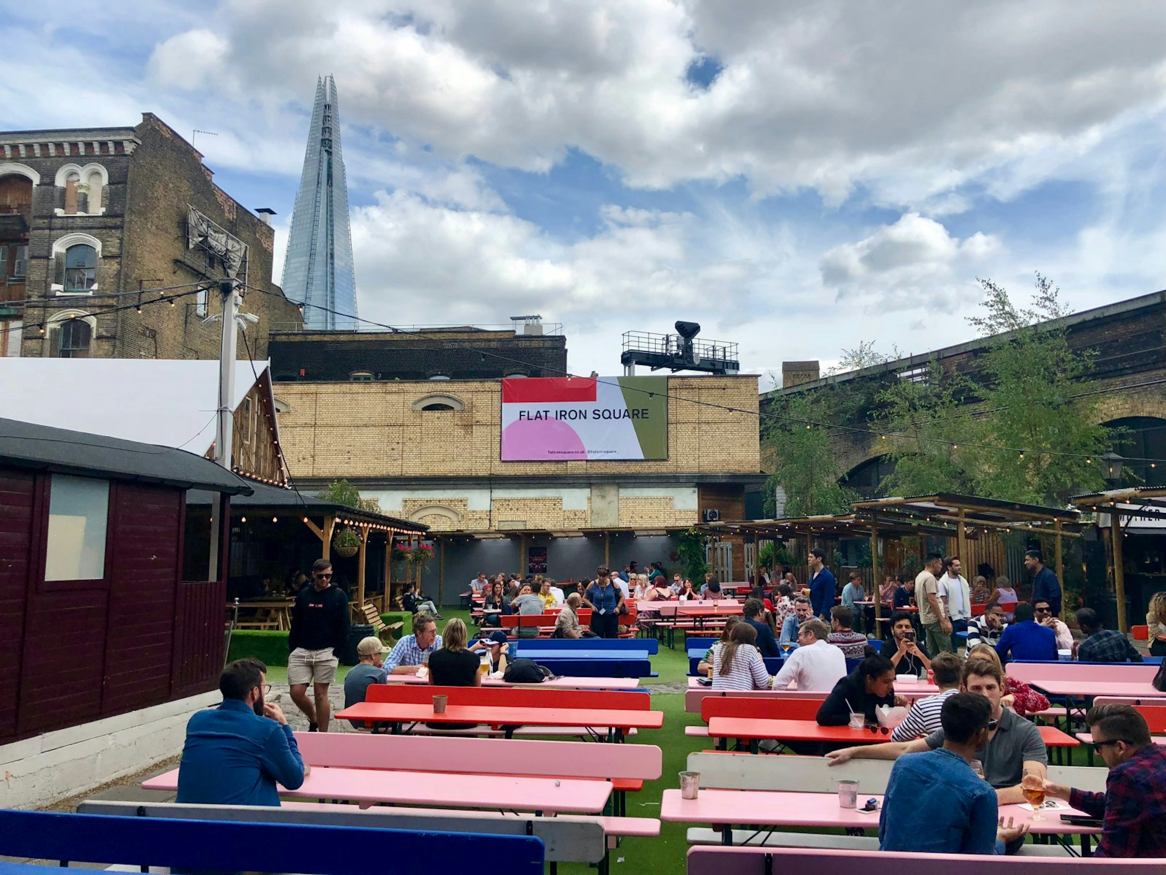 Several diners are sat at colourful benches in the outside area at Flat Iron Square. The Shard can be seen in the background. London's food halls and markets