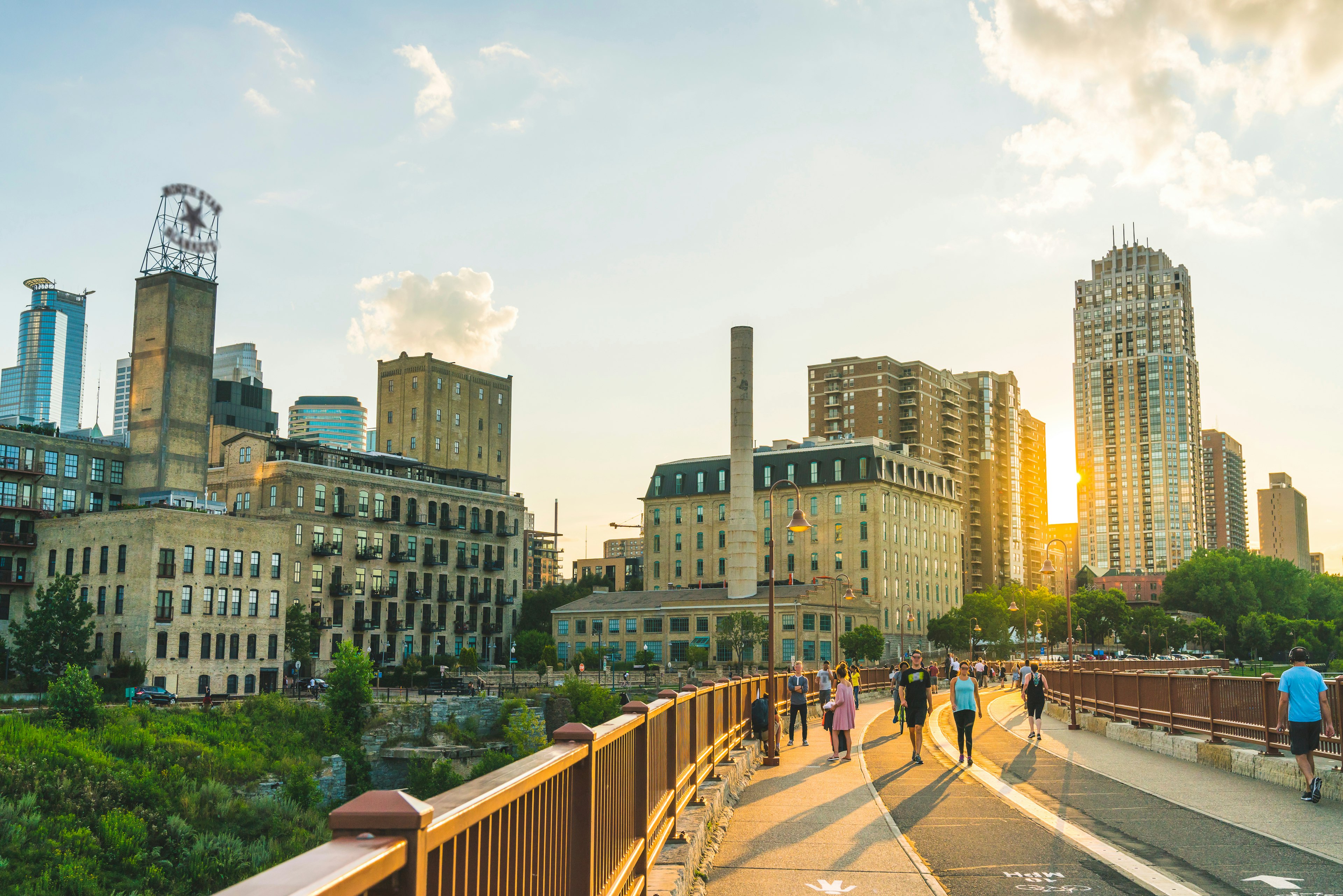 Pedestrians cross a bridge towards a series of tall buildings as the sun sets