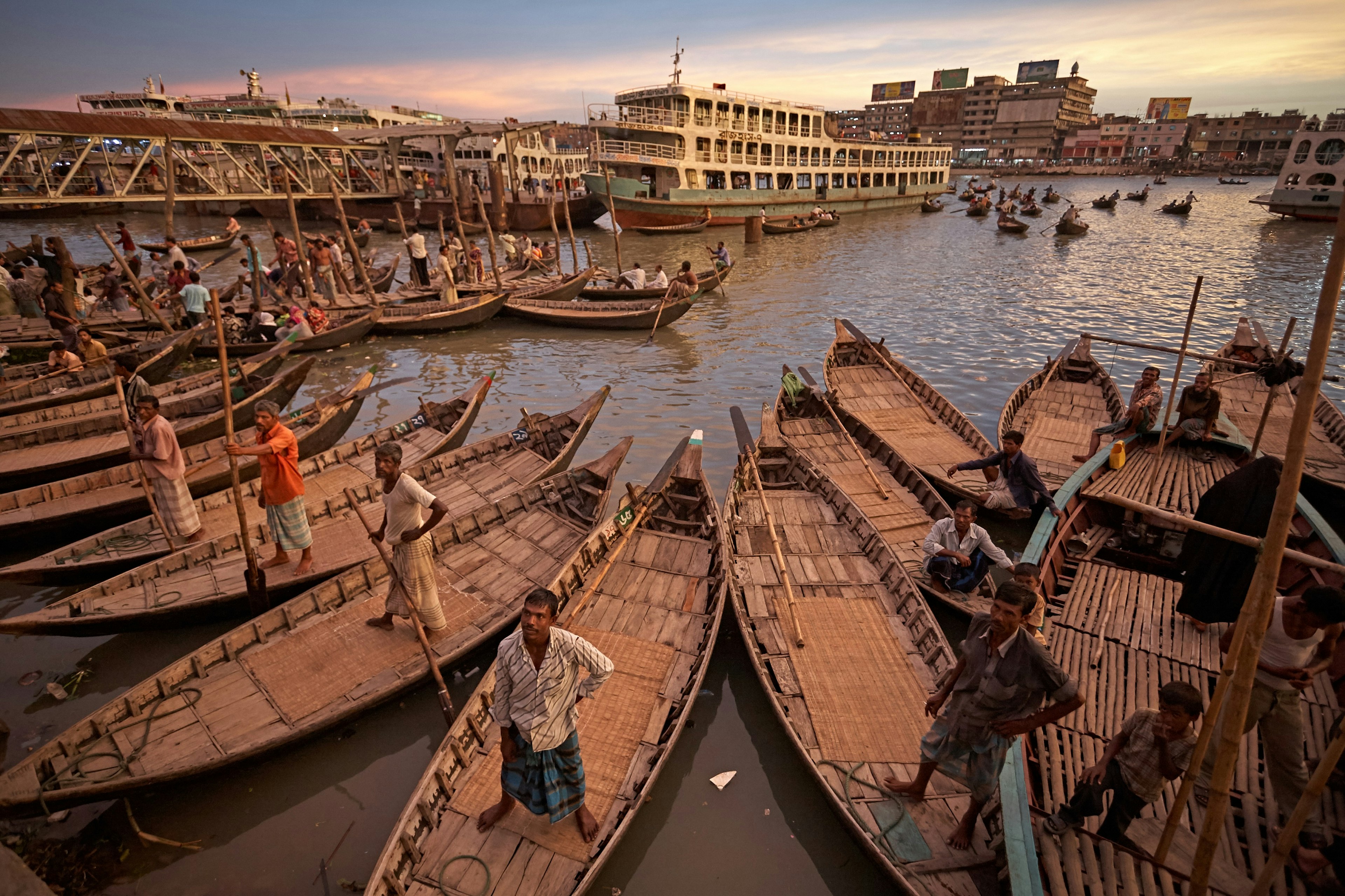 A view of the river from Dhaka's ferry terminal of Sadarghat. Many people stand in their small wooden boats, looking up towards the dock and waiting for customers. Behind, larger, passenger-cruiser style boats are visible.