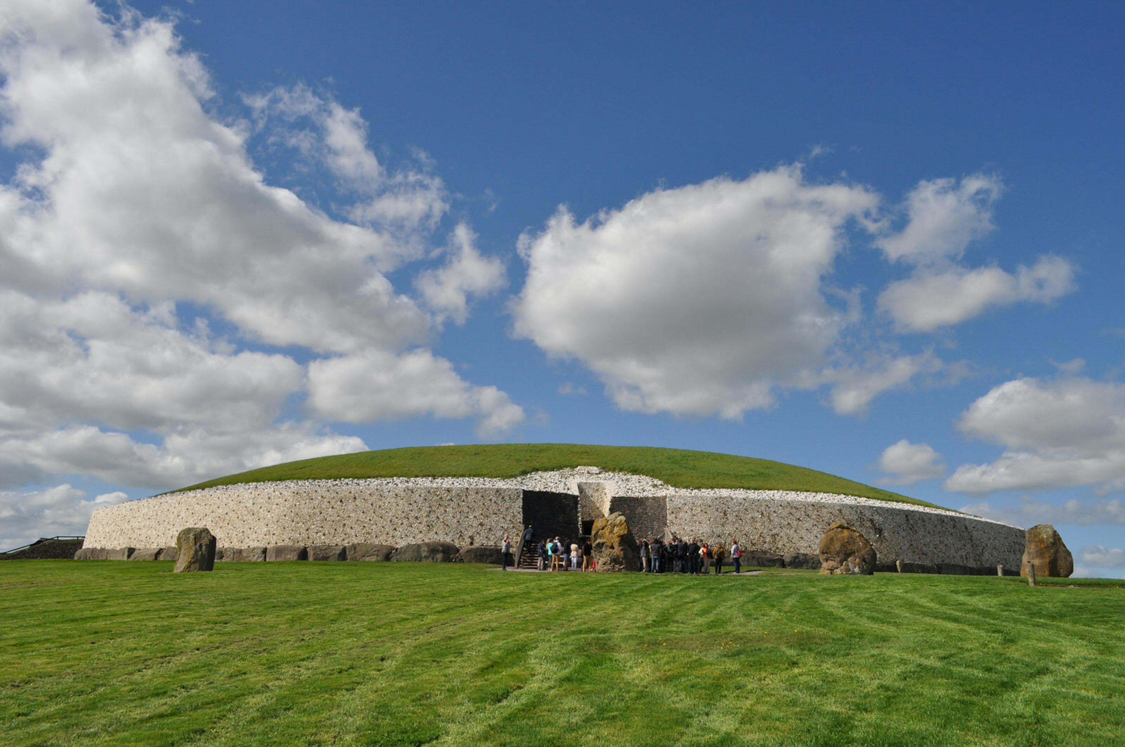 Newgrange, a prehistoric monument with a passage grave in Meath, Ireland. It is a clear day with a few fluffy clouds dotted in the blue sky.