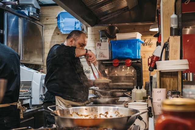 A man dressed in black standing in front of several pans on the hob serves up a plate of food