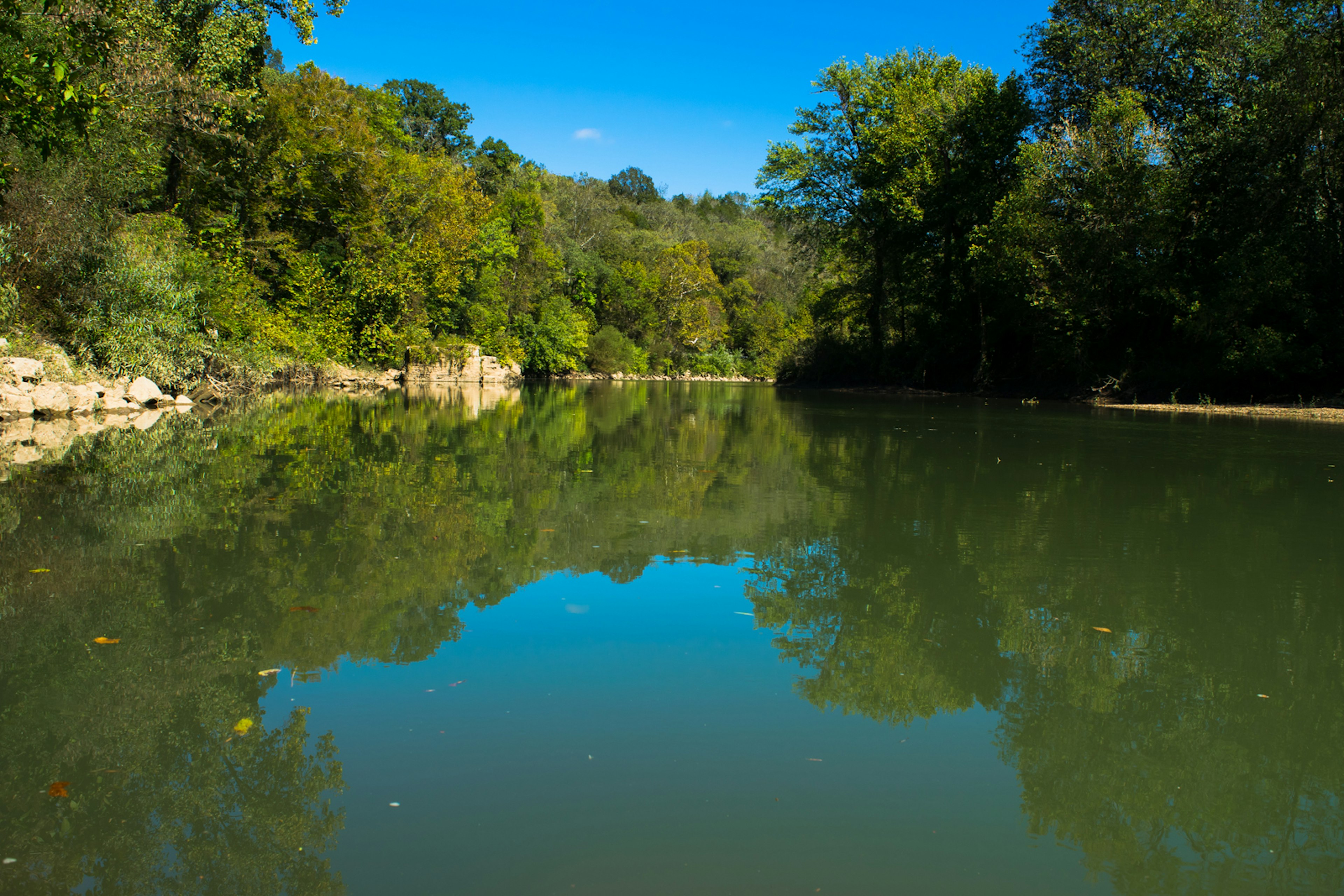 A blue sky is reflected in calm water, with trees on either side of the river; best summer trips in the Southern US