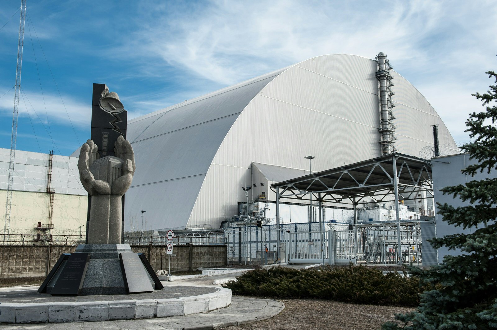 In the foreground is a concrete memorial to the Chernobyl disaster, depicting two hands cupped together around a miniature of Reactor 4; behind the memorial is the huge grey arch of the New Safe Confinement structure that encloses the remains of Reactor 4.