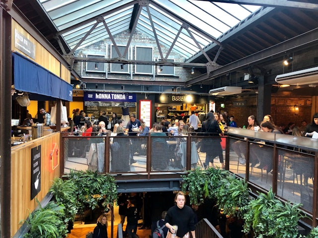 Several diners sit at tables below a glass roof in Victoria's Market Hall