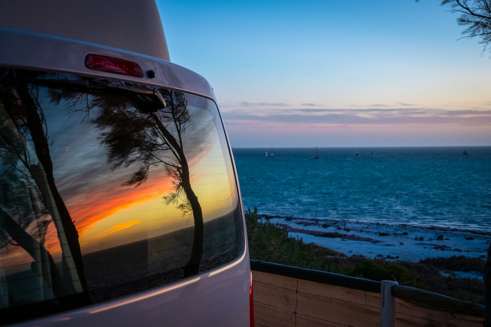 A camper van parked near the sea at sunset, with the sky and trees reflected in its rear window