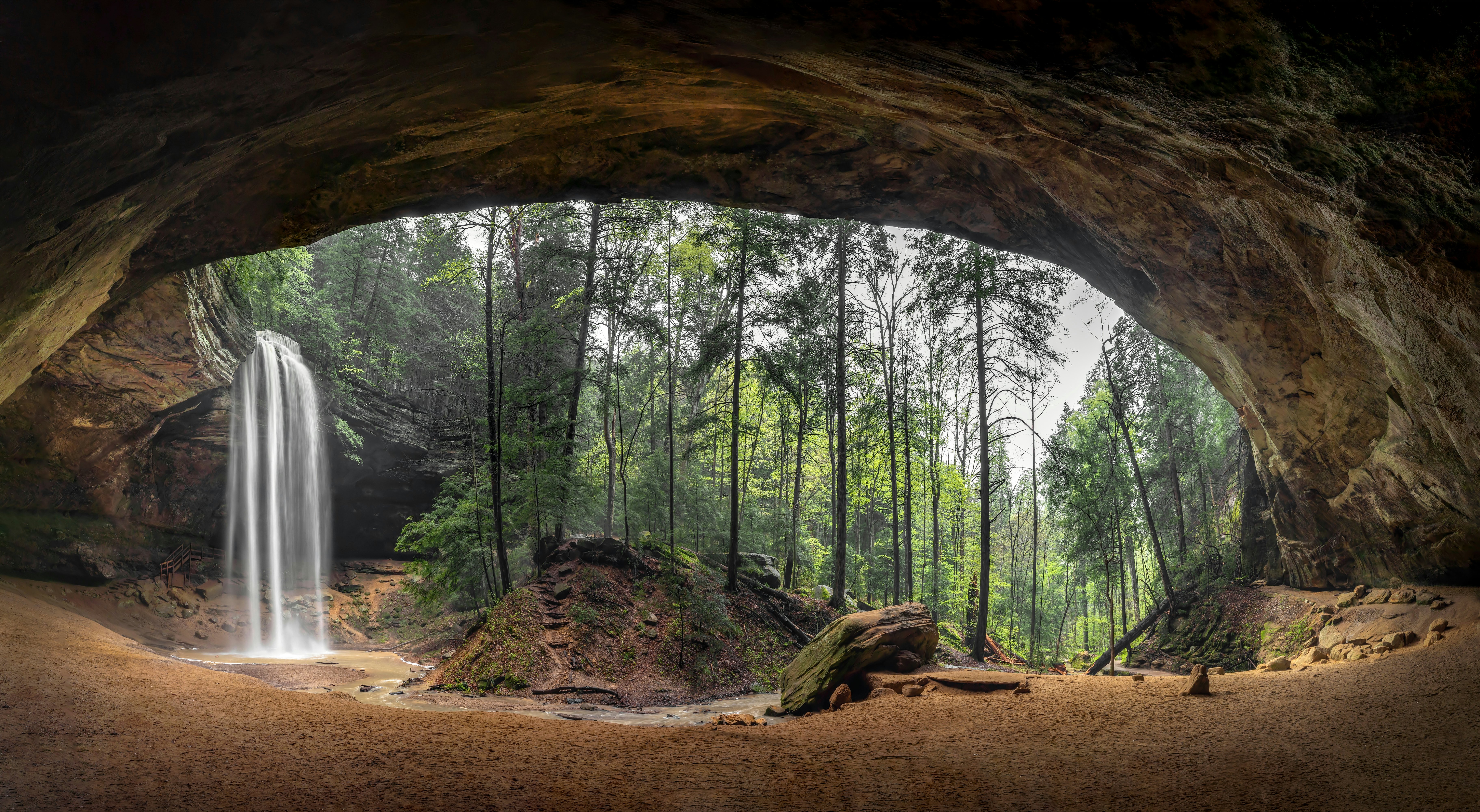 A vast cave mouth facing a cascading waterfall in woodland covered in fall foliage