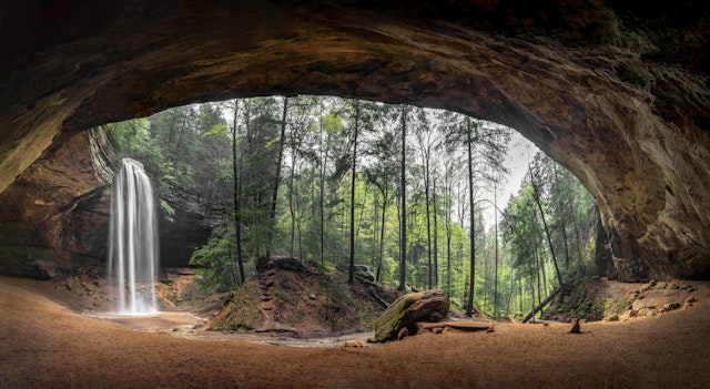 A vast cave mouth facing a cascading waterfall in woodland covered in fall foliage