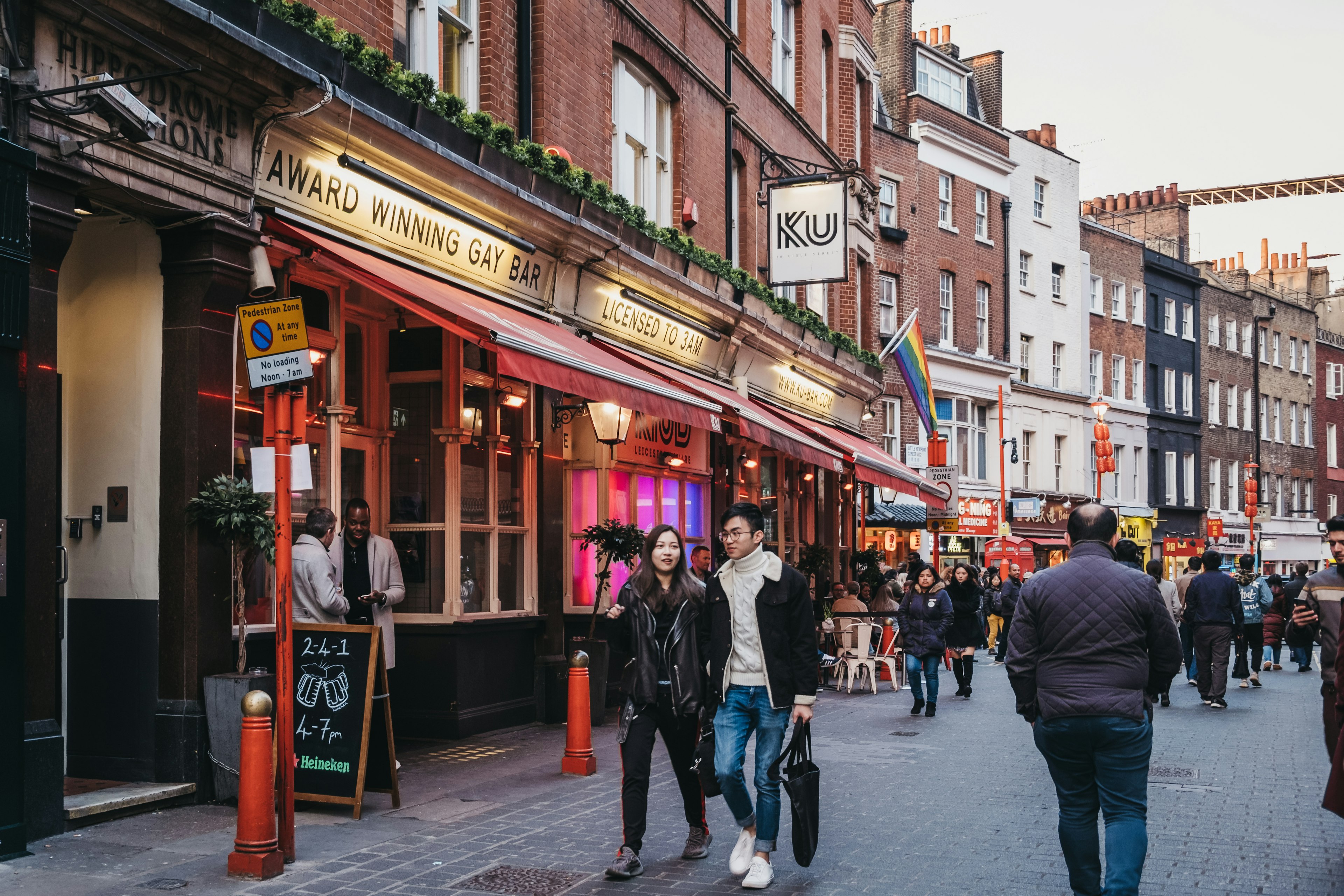 People in jackets walking past Soho's Ku Bar during the day; the bar has cream paintwork, a red awning and a line of potted plants above the windows.