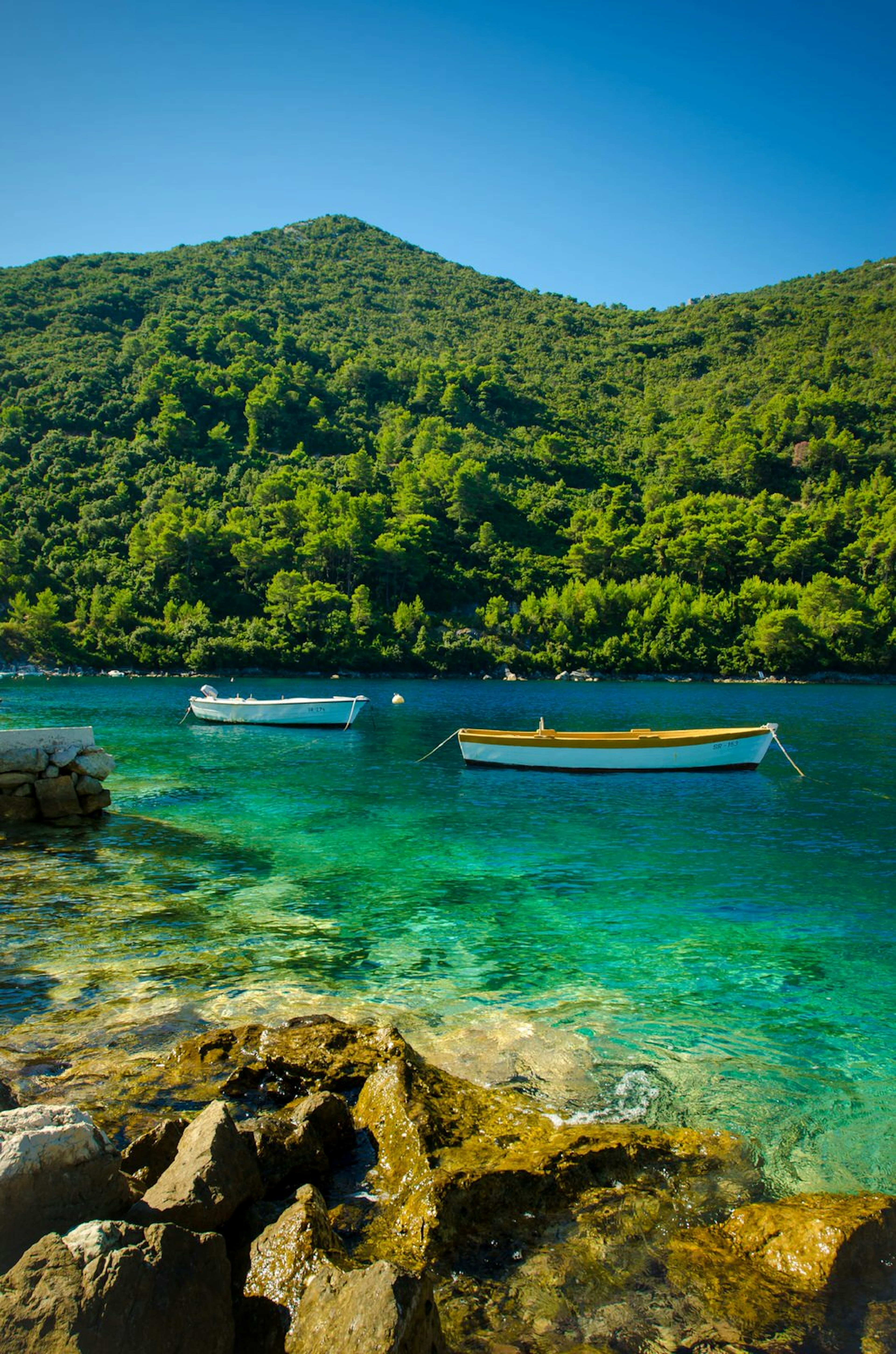 Two boats sit moored in the idyllic turquoise water of Mljet National Park; there are rocks in the foreground and forest-clad hills on the other side of the water.