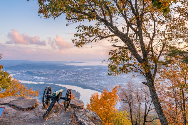 An old-style canon sits on a cliff edge pointing towards a city on the curve of a river. All the leaves on the trees in the surrounding state park are in reds and golds of autumn