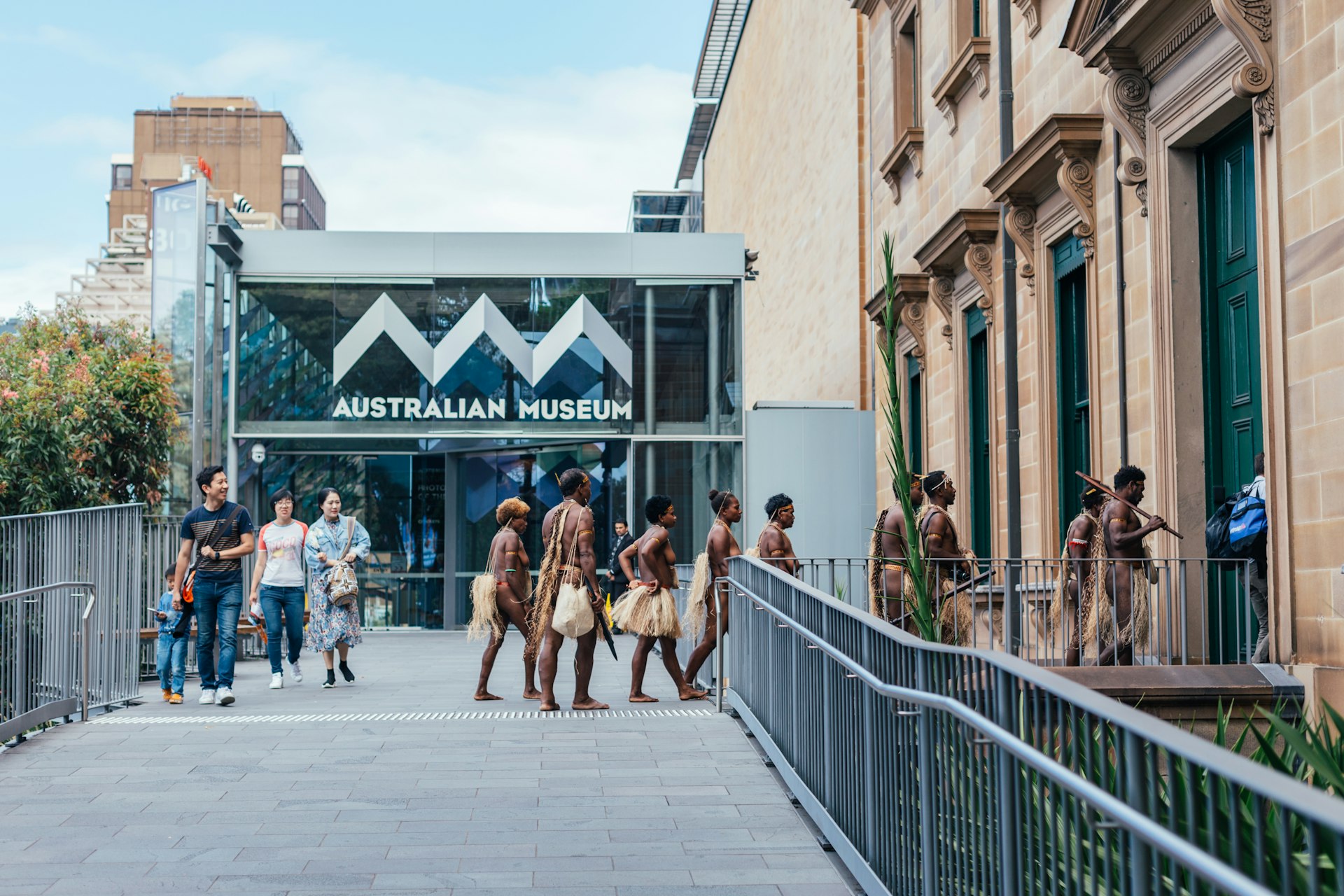 The exterior of the Australian Museum building with a group of Aboriginal people entering barefoot wearing traditional skirts