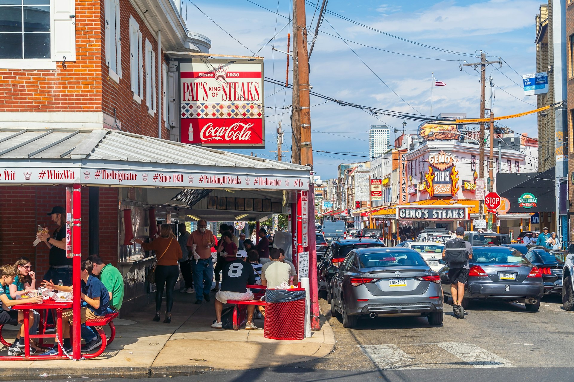 A busy street intersection with a Pat's King of Steaks diner on one side and Geno's Steaks further down the road