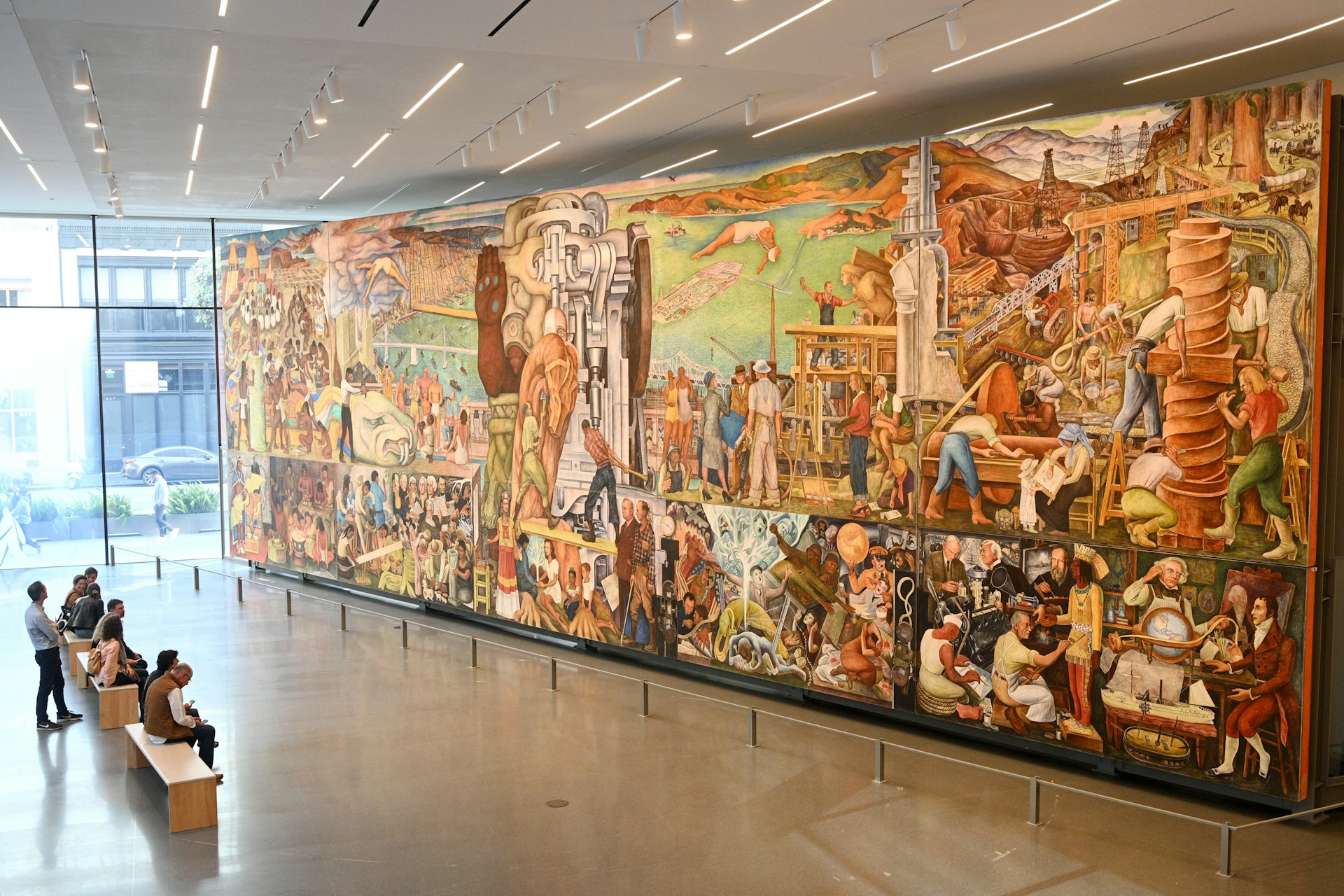 People sit on a bench in a large gallery gazing at a mural that takes up one side of the room