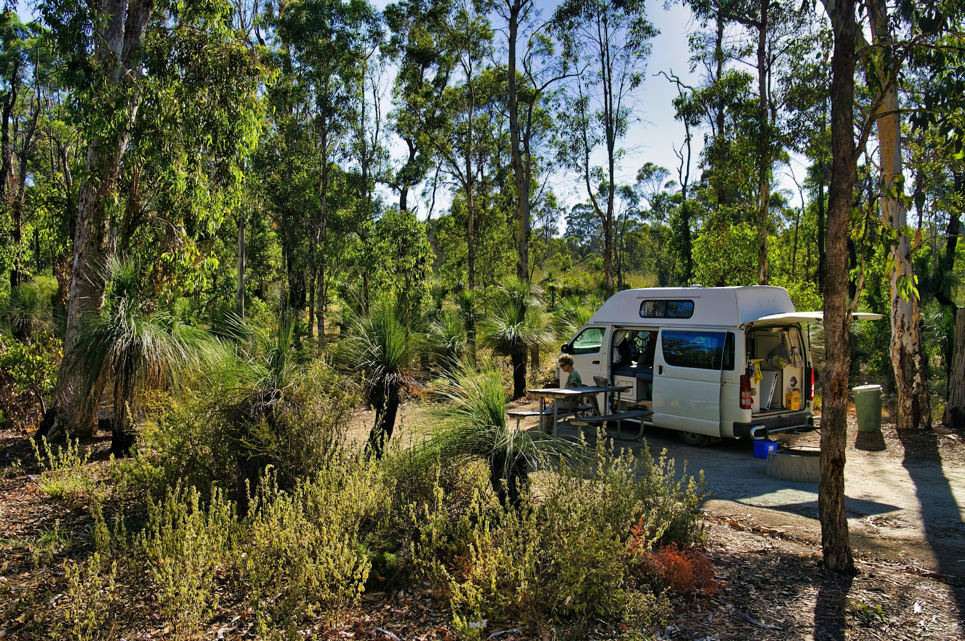 A camper van parked in a woodland camp site with doors open, tables out and people sat nearby