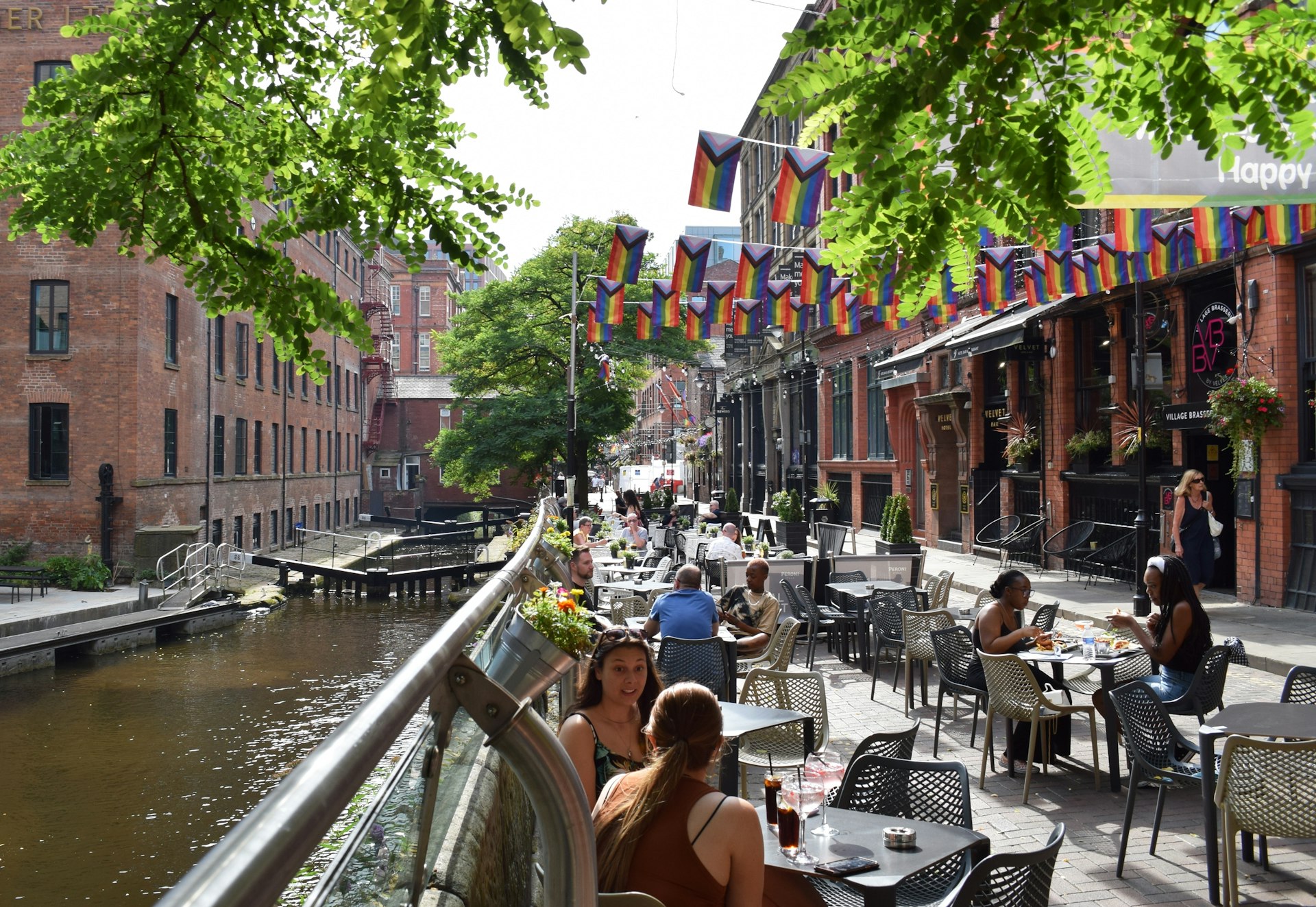 People sit at tables under rainbow flags along Canal St by the Rochdale Canal, in the heart of the Gay Village in Manchester, England