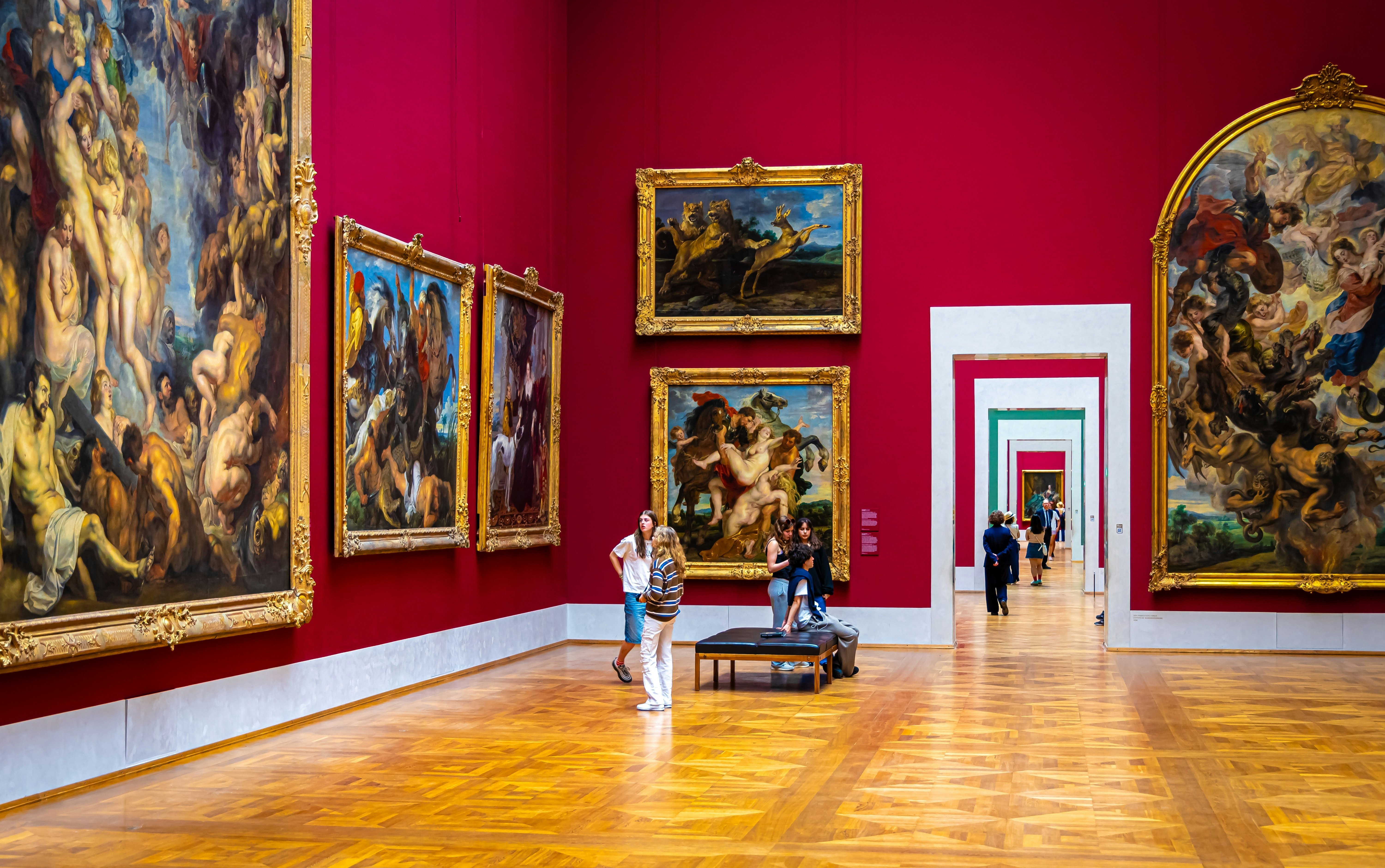 People walk through galleries with red walls hung with huge old master paintings at the Alte Pinakothek museum, Munich