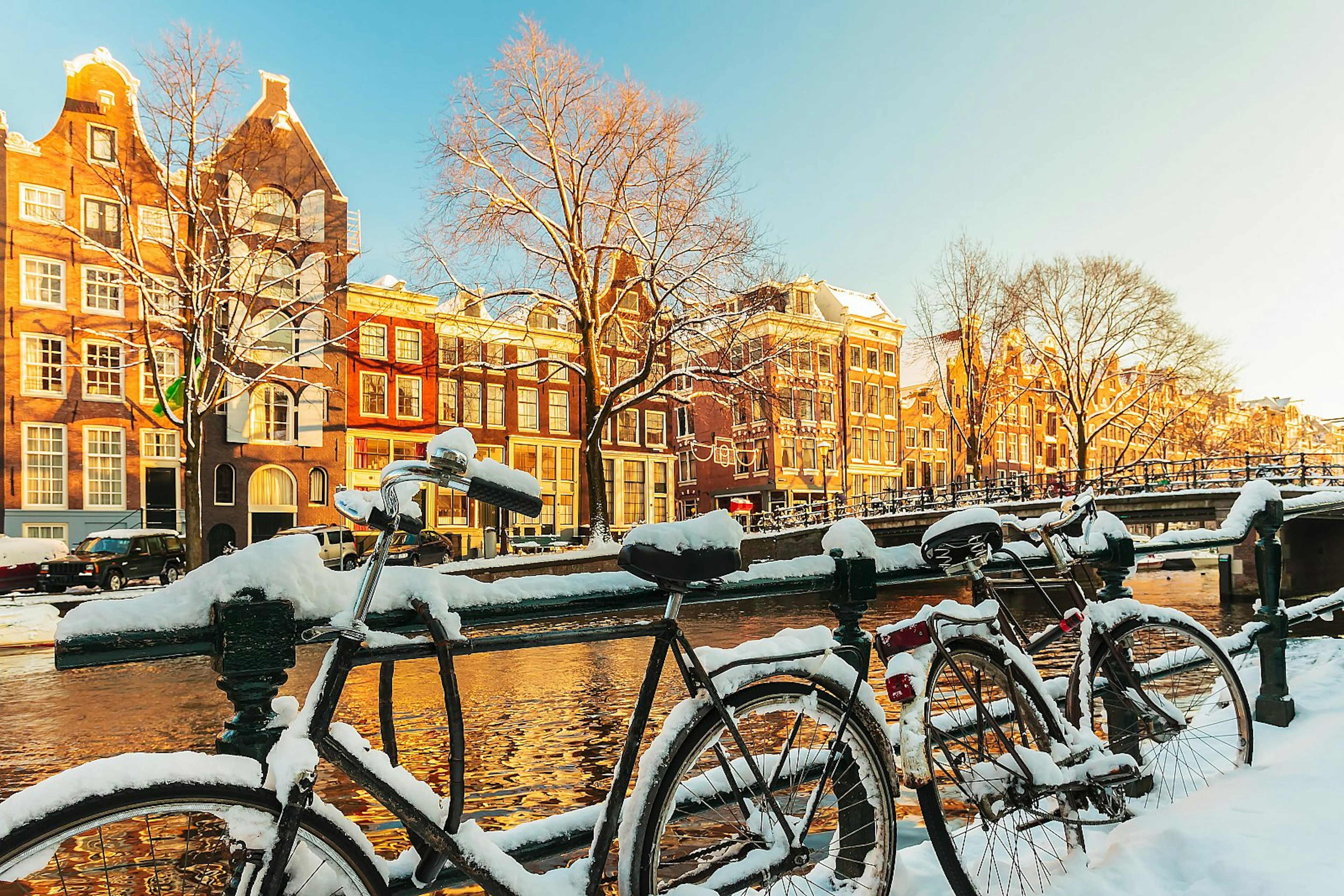 Snow-covered bicycles resting on a railing with an Amsterdam canal behind it; on the other side of the canal are gabled townhouses. Amsterdam winter