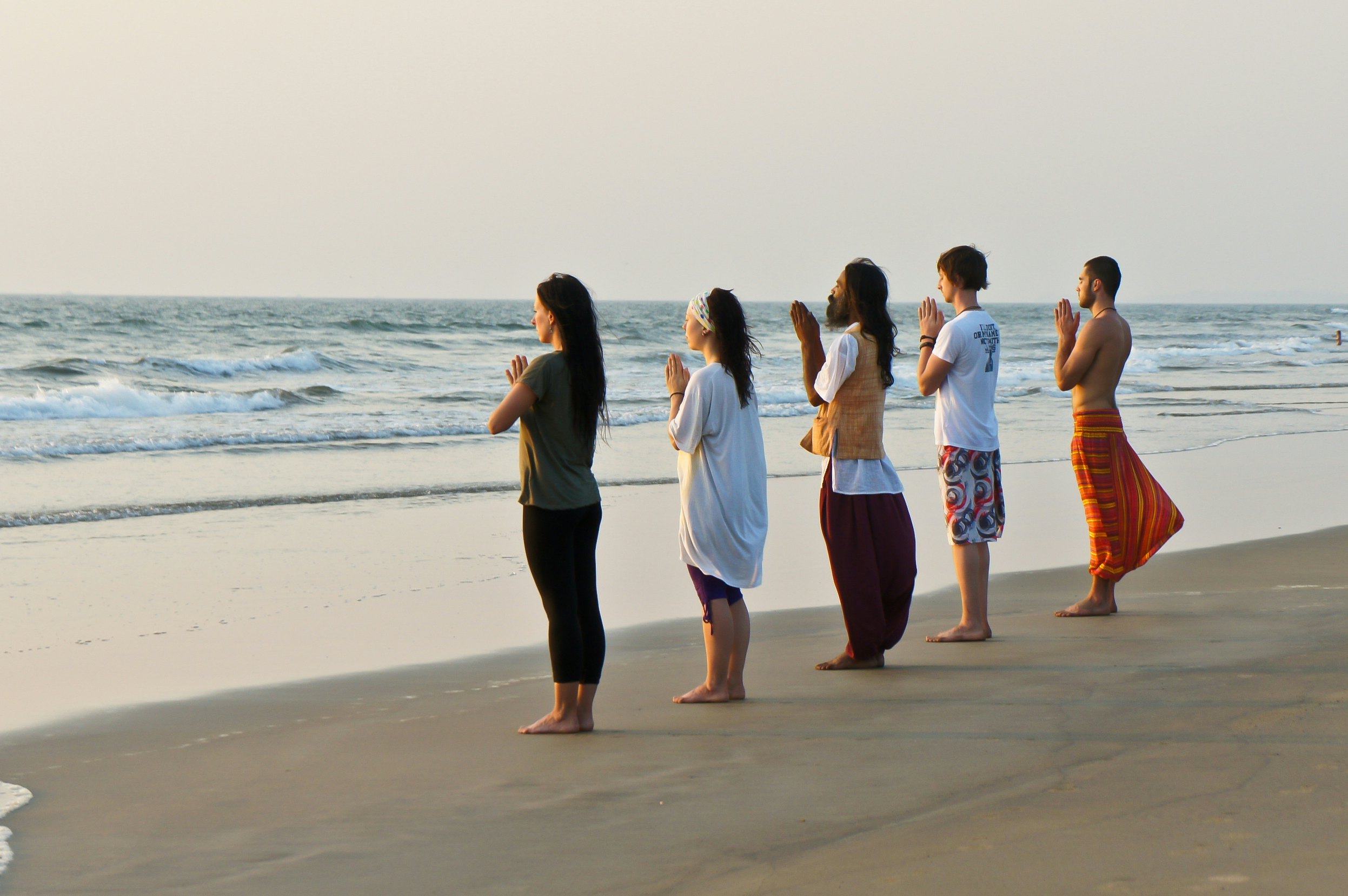 Five people stand side by side facing the ocean with their hands clasped as if in prayer as they meditate.