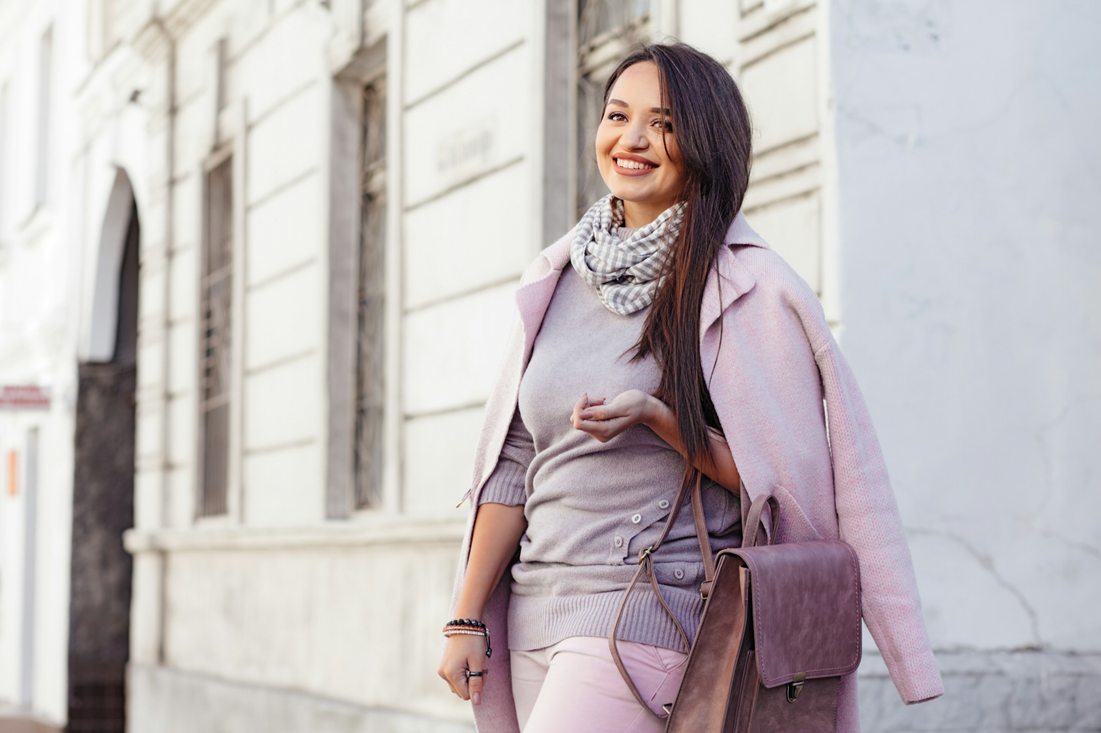 Young stylish woman wearing pink warm coat across her shoulders, light-coloured pants and handbag walking in the city street in cold season.