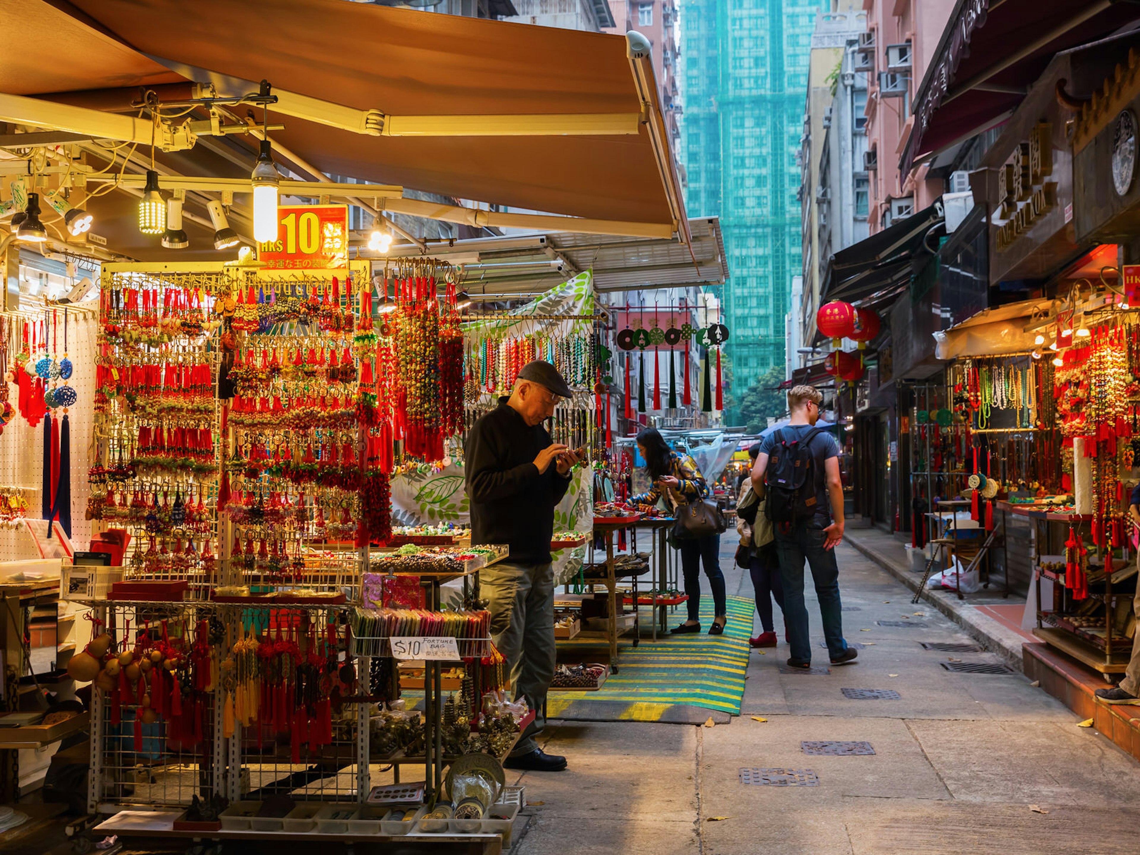 A stall with awning lit by bare bulbs selling red tassels and temple trinkets on a small Hong Kong side street