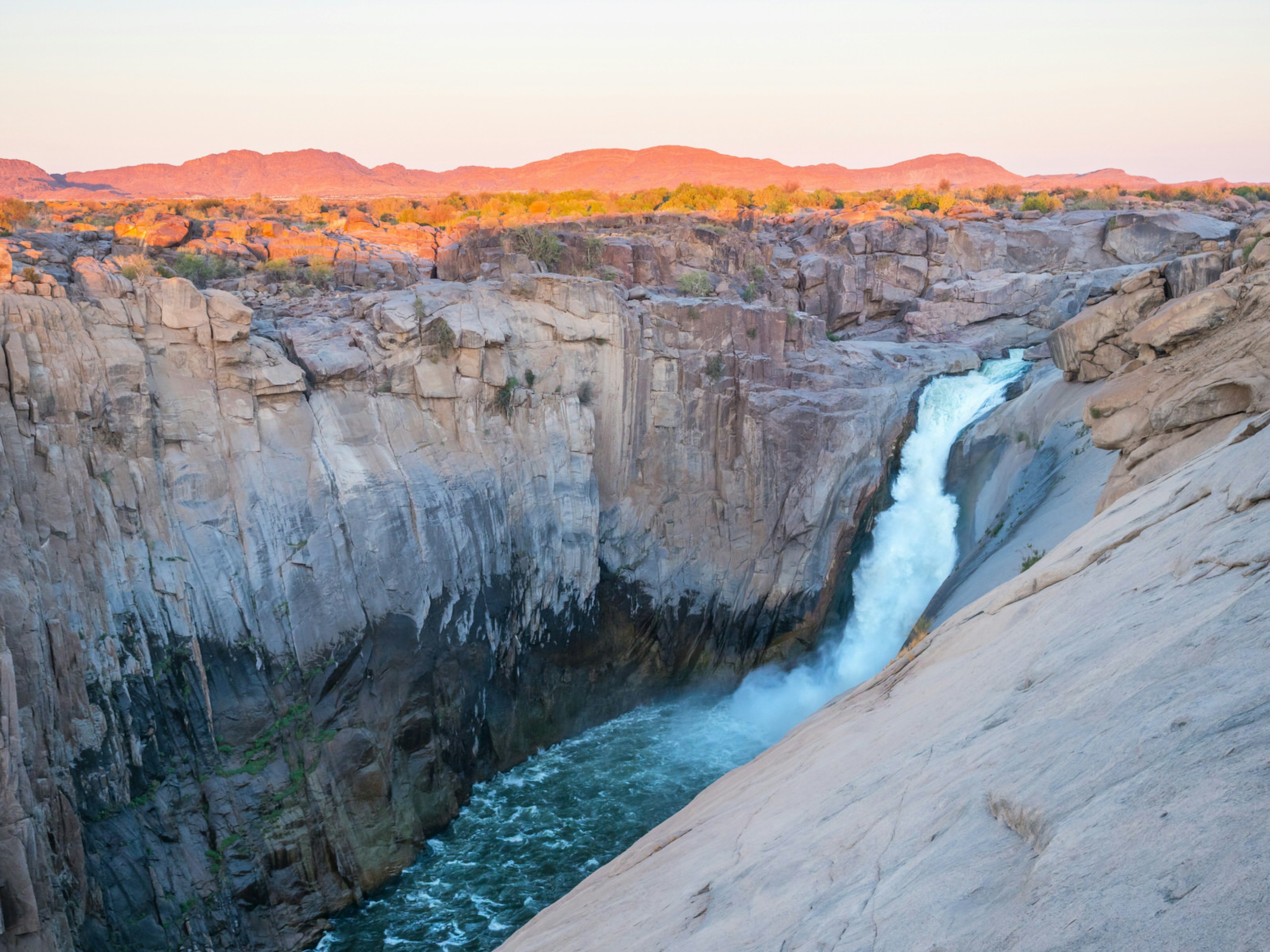 A waterfall plunges into a deep canyon, with orange hills in the background