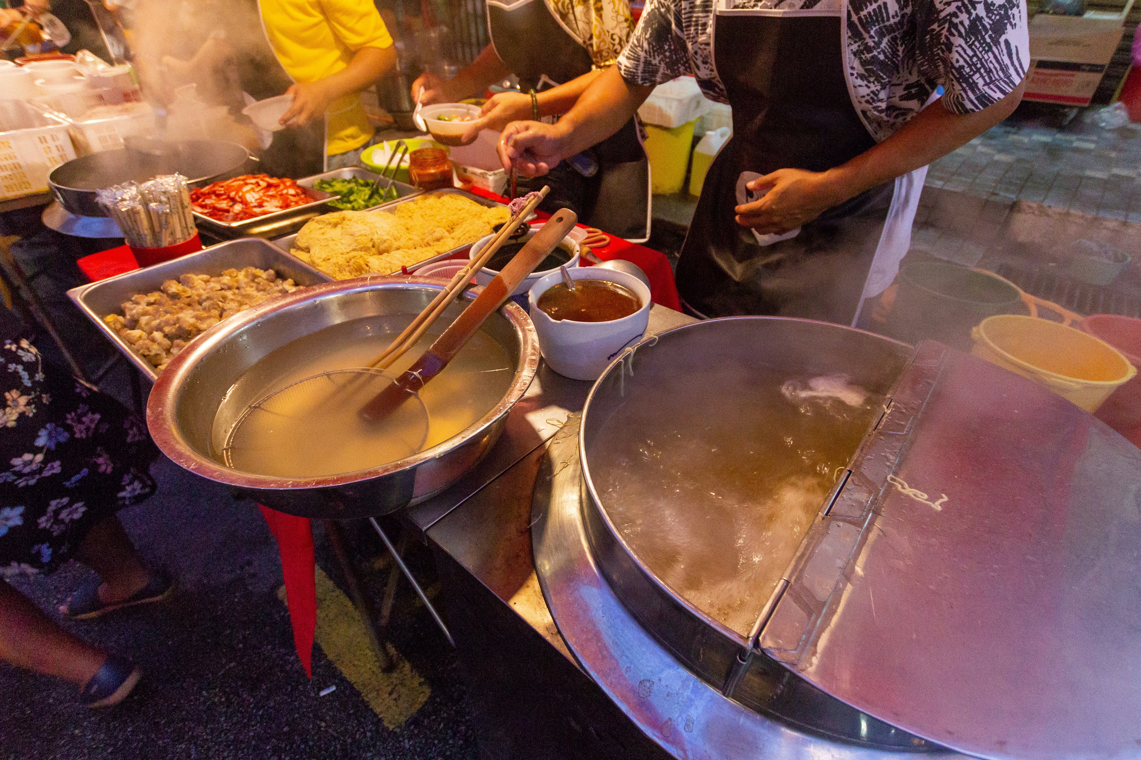 Looking down on a street food stall. There is a huge, stainless-steel vat of boiling water, and the vendors are only visible from the torso down; they are spooning various ingredients from different containers.