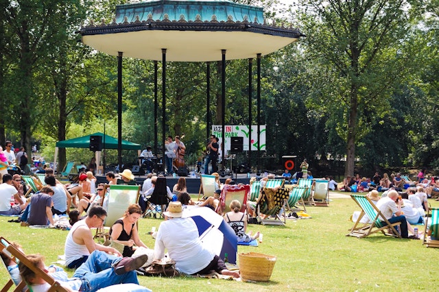 People sitting in a London park in the sunshine, while a jazz band performs in a bandstand.