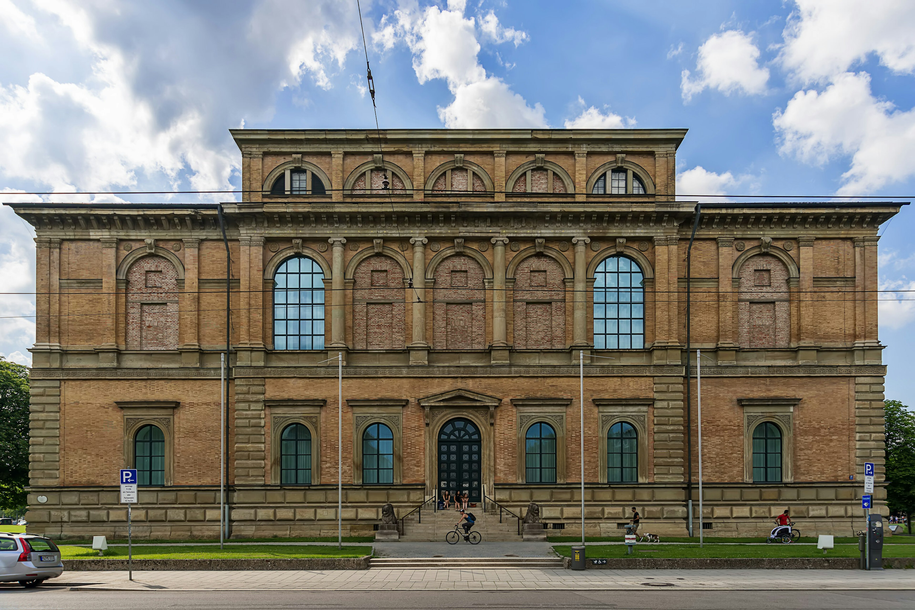 Brick, neoclassical-style building with arched windows and a large black arched door with steps leading up to it. Some people are sitting on the steps and two cyclists are riding past.